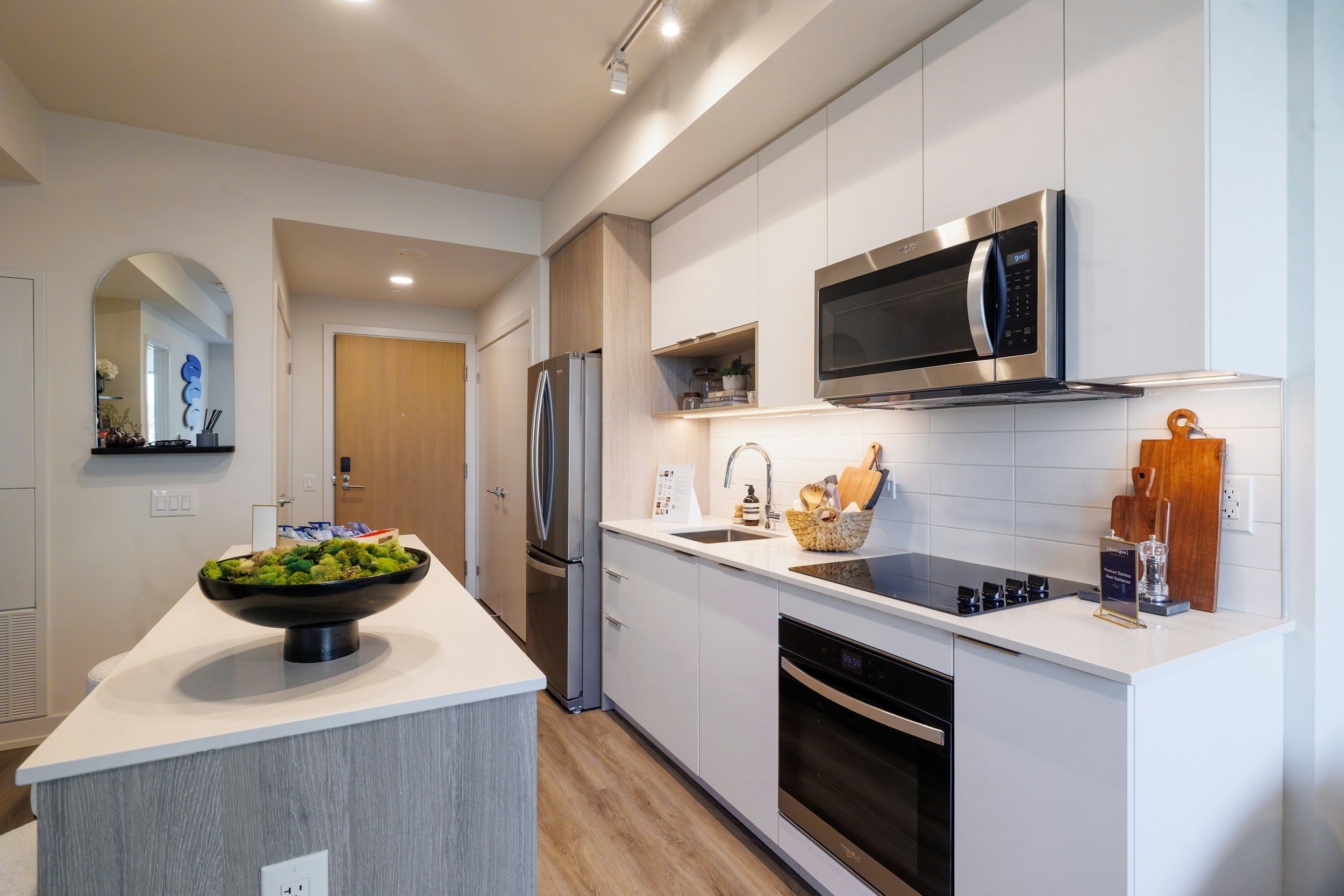 Modern kitchen with white cabinets, stainless steel microwave, black oven, refrigerator, sink, cutting boards, and decorative items. There is an island with a bowl of plants and a doorway leading to other rooms.