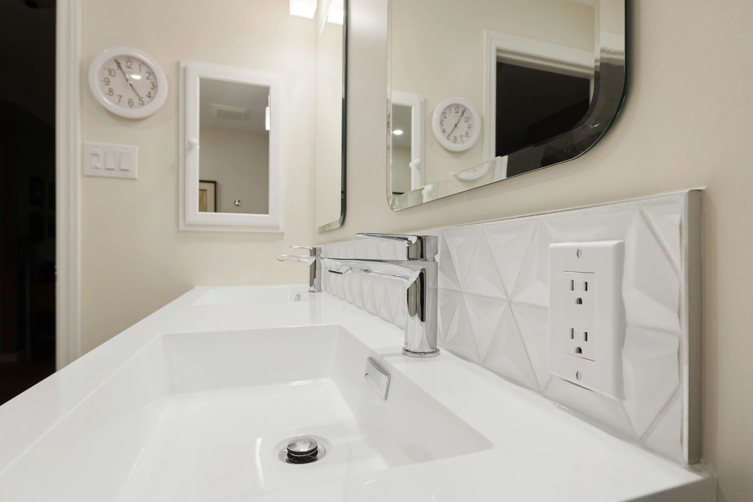 Close-up view of a white bathroom vanity with a rectangular sink, modern chrome faucet, mirror above the sink, and a wall clock showing 10:24. The wall has a switch plate and an electrical outlet.