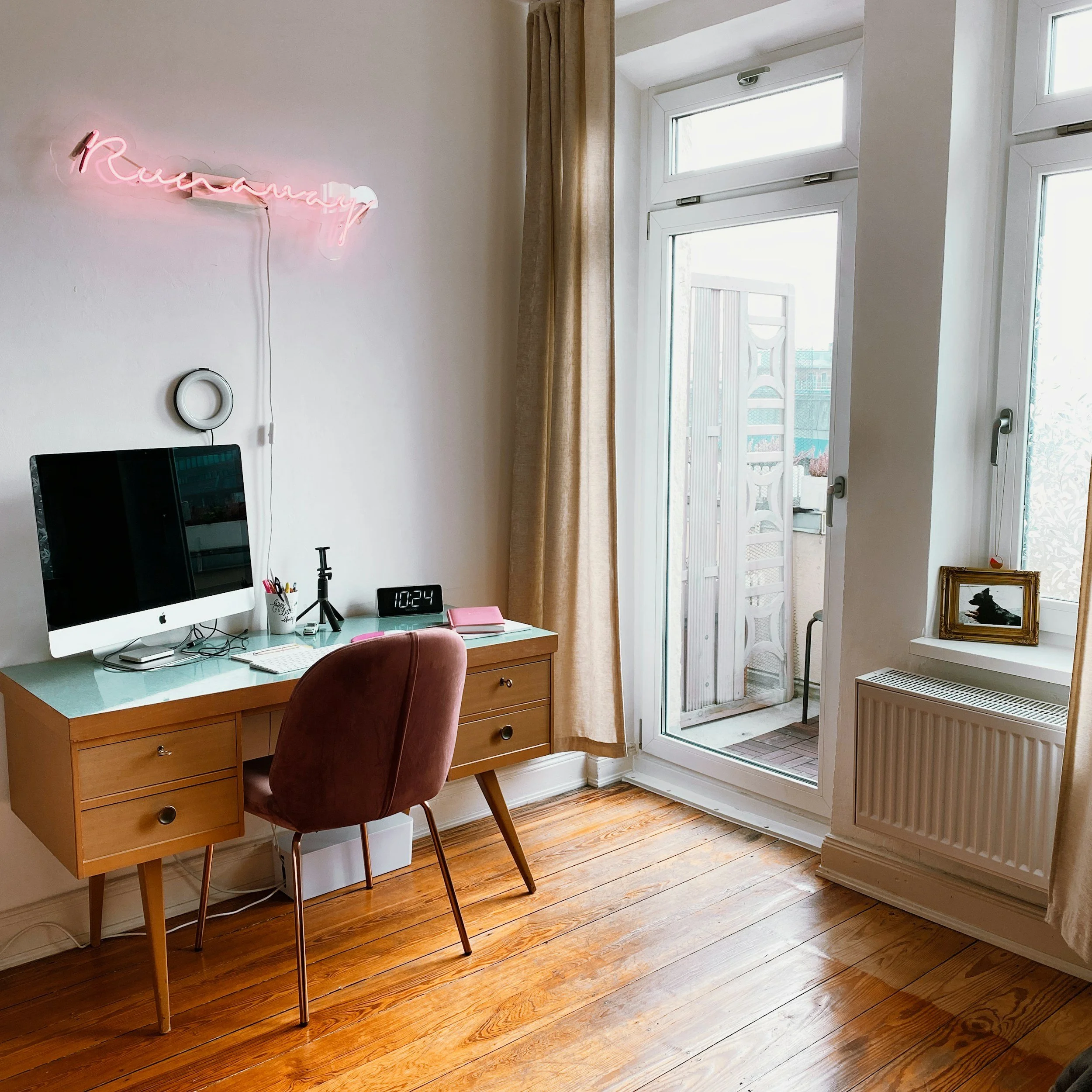A home office with a wooden desk, an iMac computer, a pink notebook, a coffee mug, and a digital clock showing 10:24. The wall has a pink neon sign that reads "pursuing" and a white circle mirror. Large windows and a door lead to a balcony with outdoor furniture.