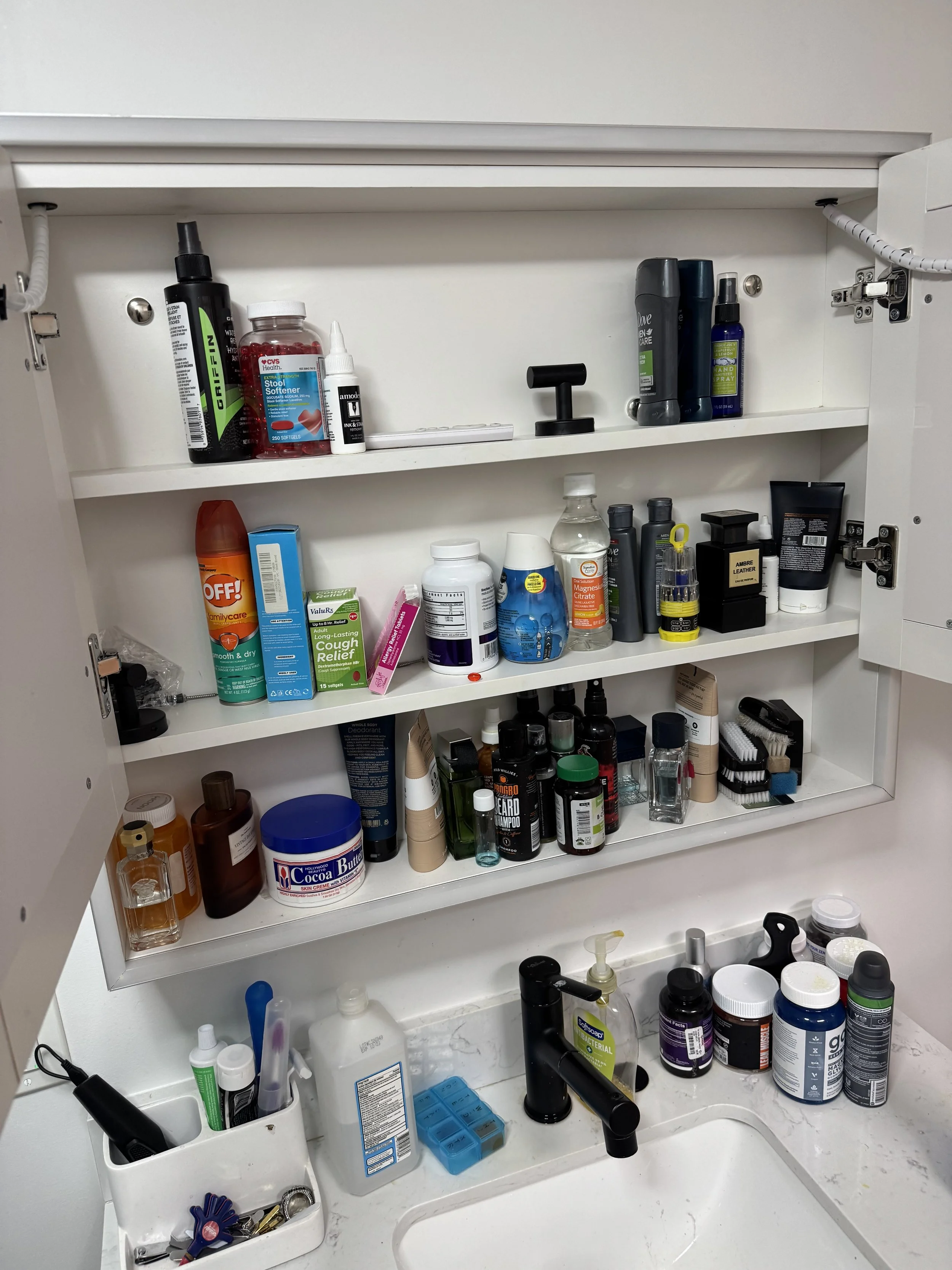 Clutterd Bathroom medicine cabinet with various personal care and health products, including ointments, vitamins, sprays, and bottles, above a sink with a soap dispenser and toothbrushes.