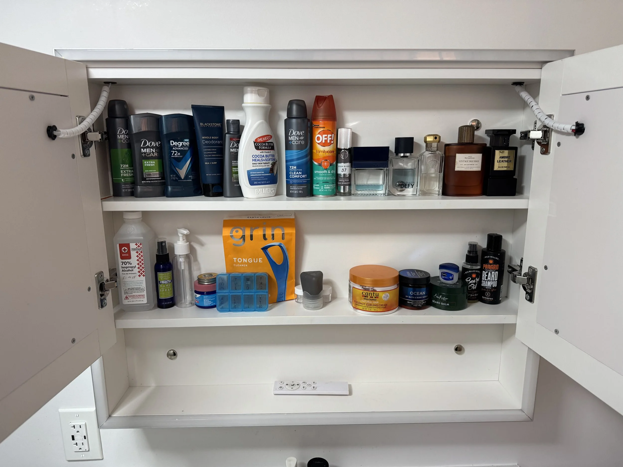 Open white bathroom cabinet with two shelves containing various personal care products like deodorant, body spray, lotion, perfumes, and grooming items. The lower shelf is mostly empty with a remote control and a few small items. The cabinet has electrical outlets and a white wall background.
