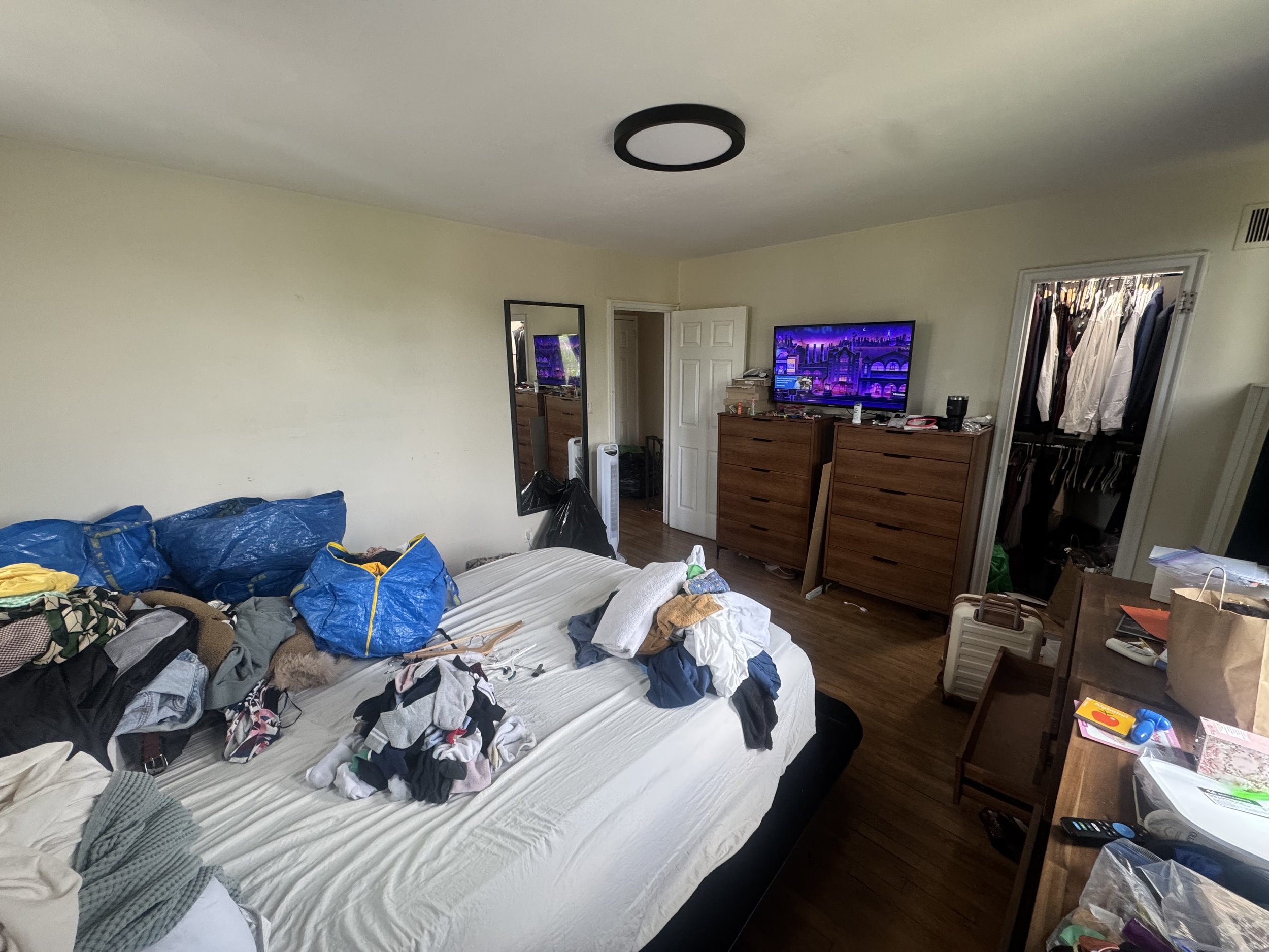 A bedroom with a disorganized bed covered with clothes, a dresser with a television, and an open closet with clothes hanging inside.