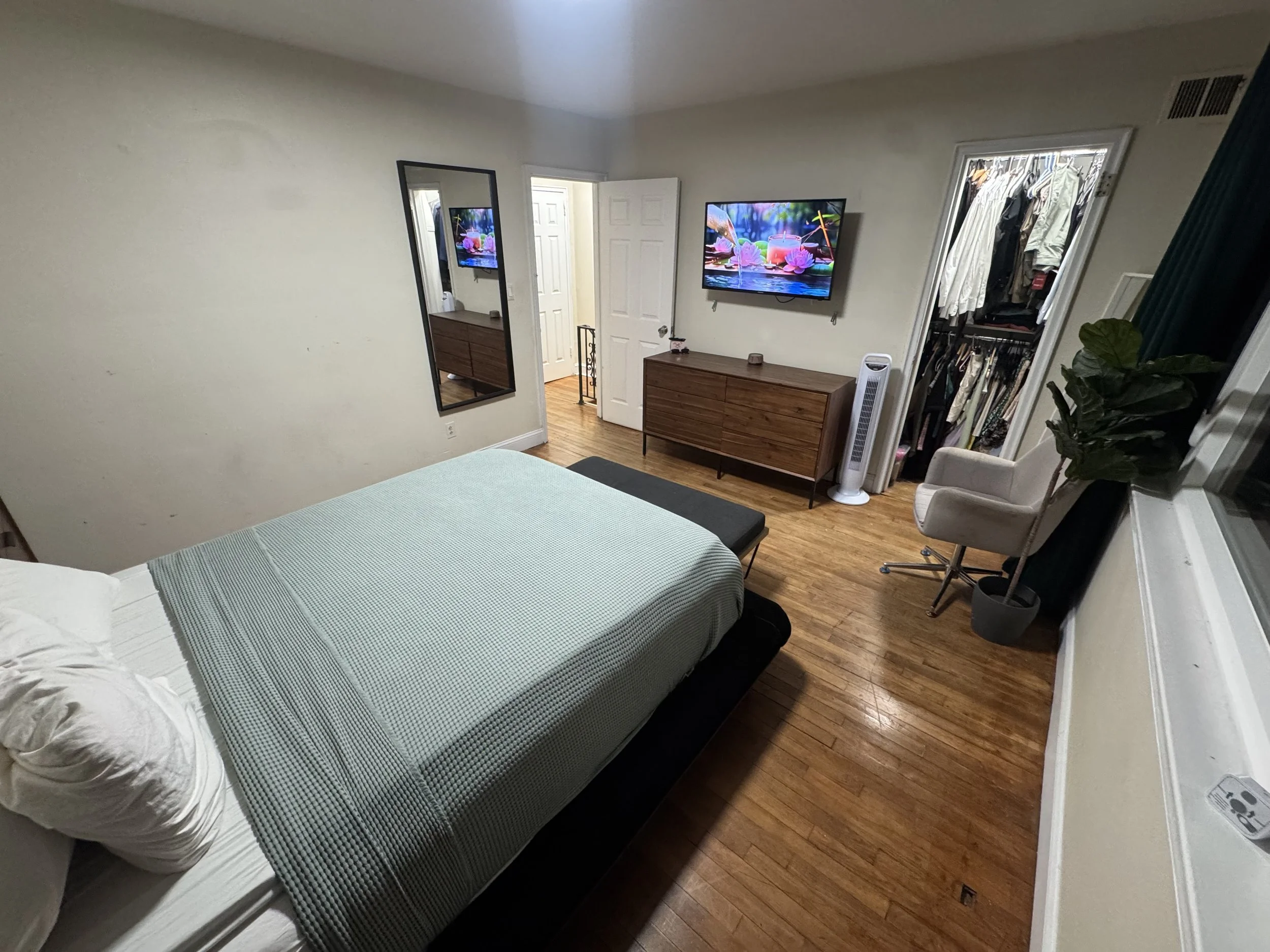 A Organized bedroom with a bed, a dresser, a wall-mounted TV showing a colorful nature scene, a mirror, an open closet with clothes, a tower fan, and a chair with a large green plant nearby.