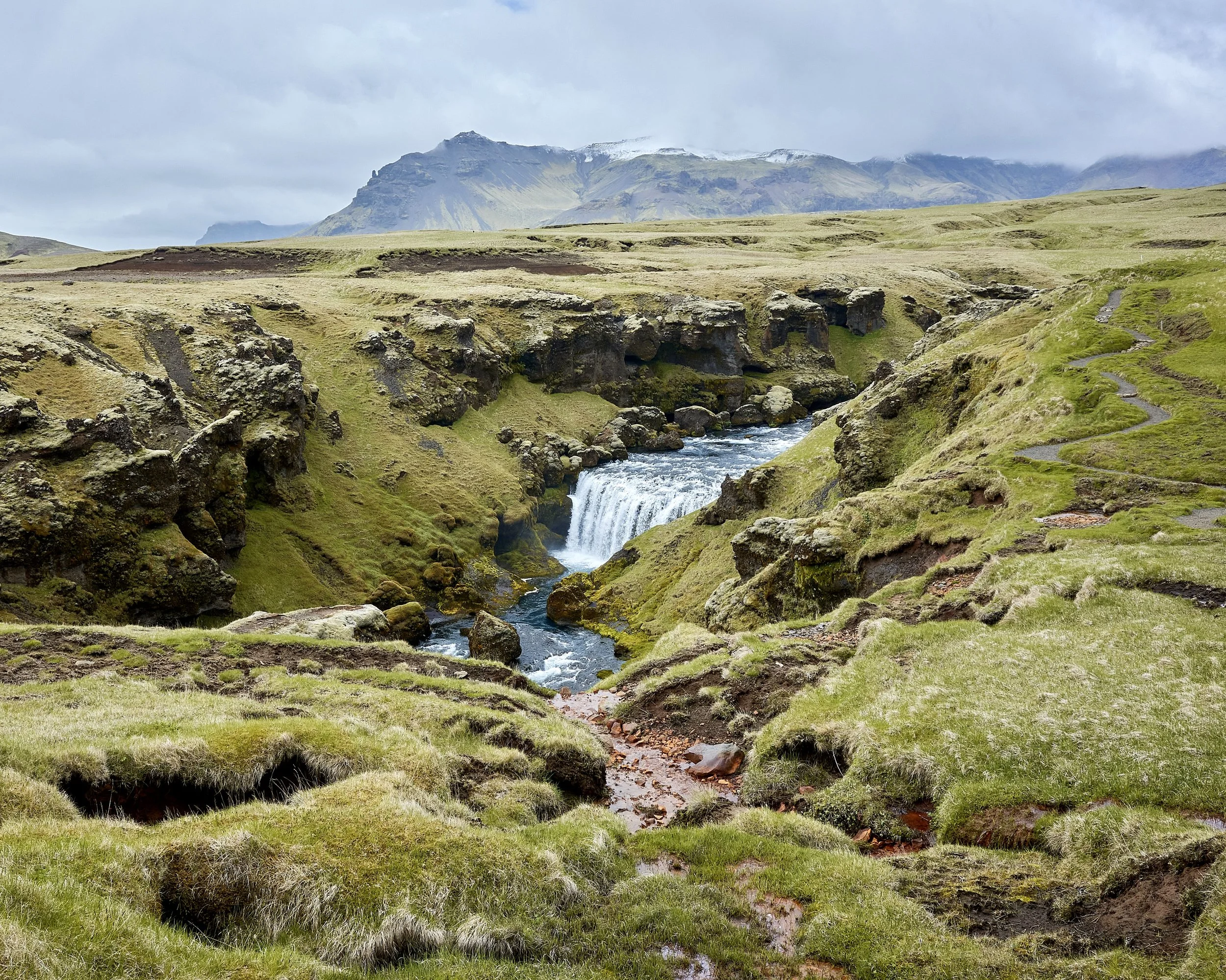 Fimmvörðuháls, Iceland. A scenic landscape of moss-covered rocky terrain with a flowing river and a waterfall, green hills, and mountains in the distance under a cloudy sky.