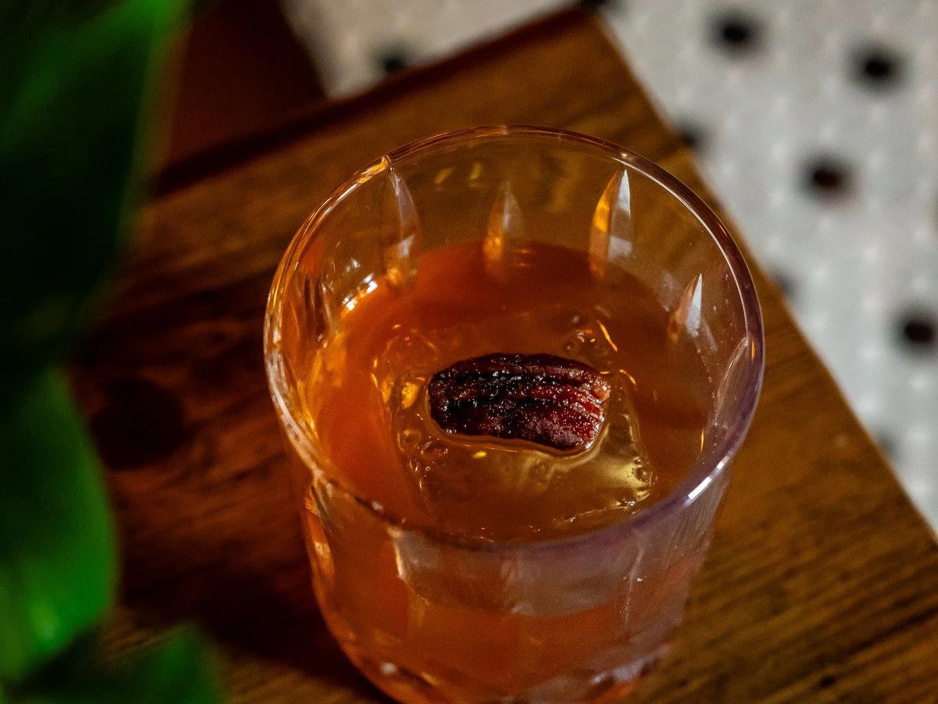 A glass of amber-colored beverage with a large ice cube and a piece of dried fruit, possibly a date, on top, placed on a wooden surface.