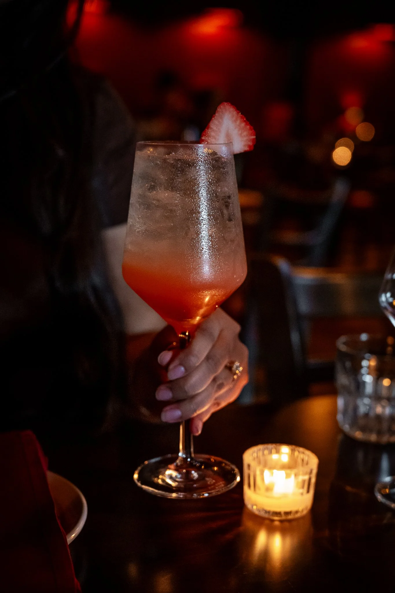 A woman holding a cocktail glass with a slice of strawberry on the rim in a dimly lit setting with a candle on the table.