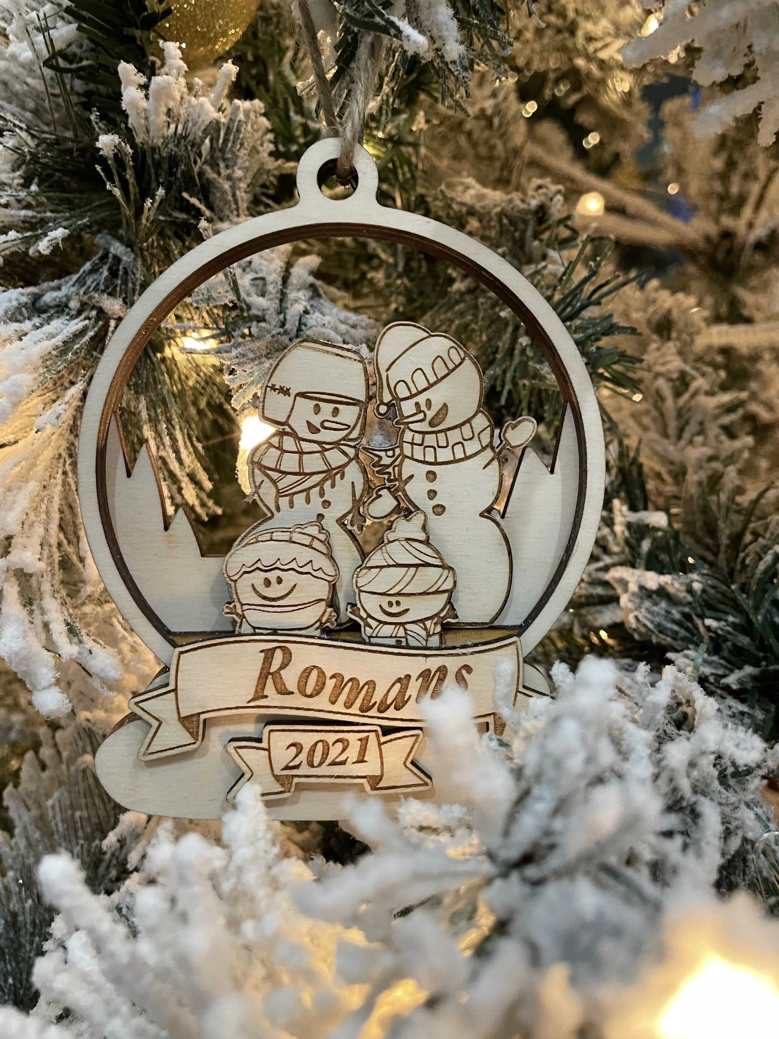 A wooden Christmas ornament featuring a snowman, two children, and a Christmas tree, with the word 'Romans' and the year '2021' inscribed on it, hanging on a snow-covered Christmas tree.