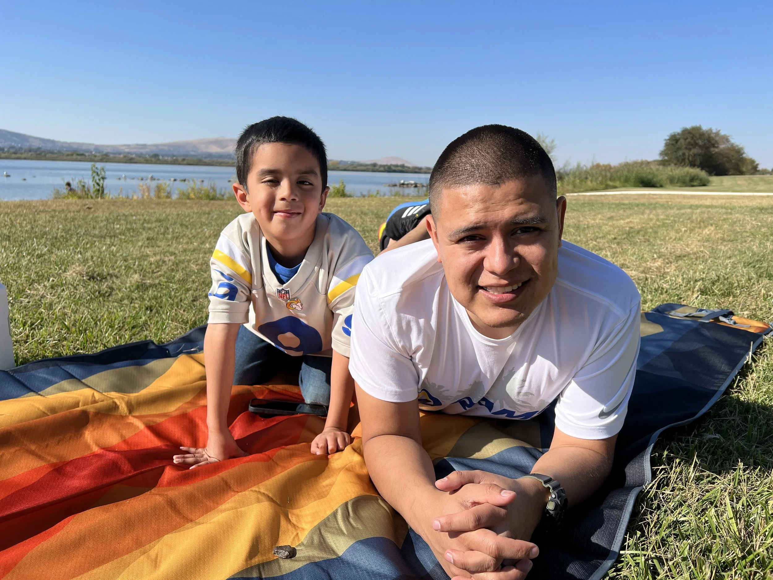 A man and a boy lying on a colorful blanket on the grass near a lake, smiling at the camera with the lake and hills in the background on a sunny day.