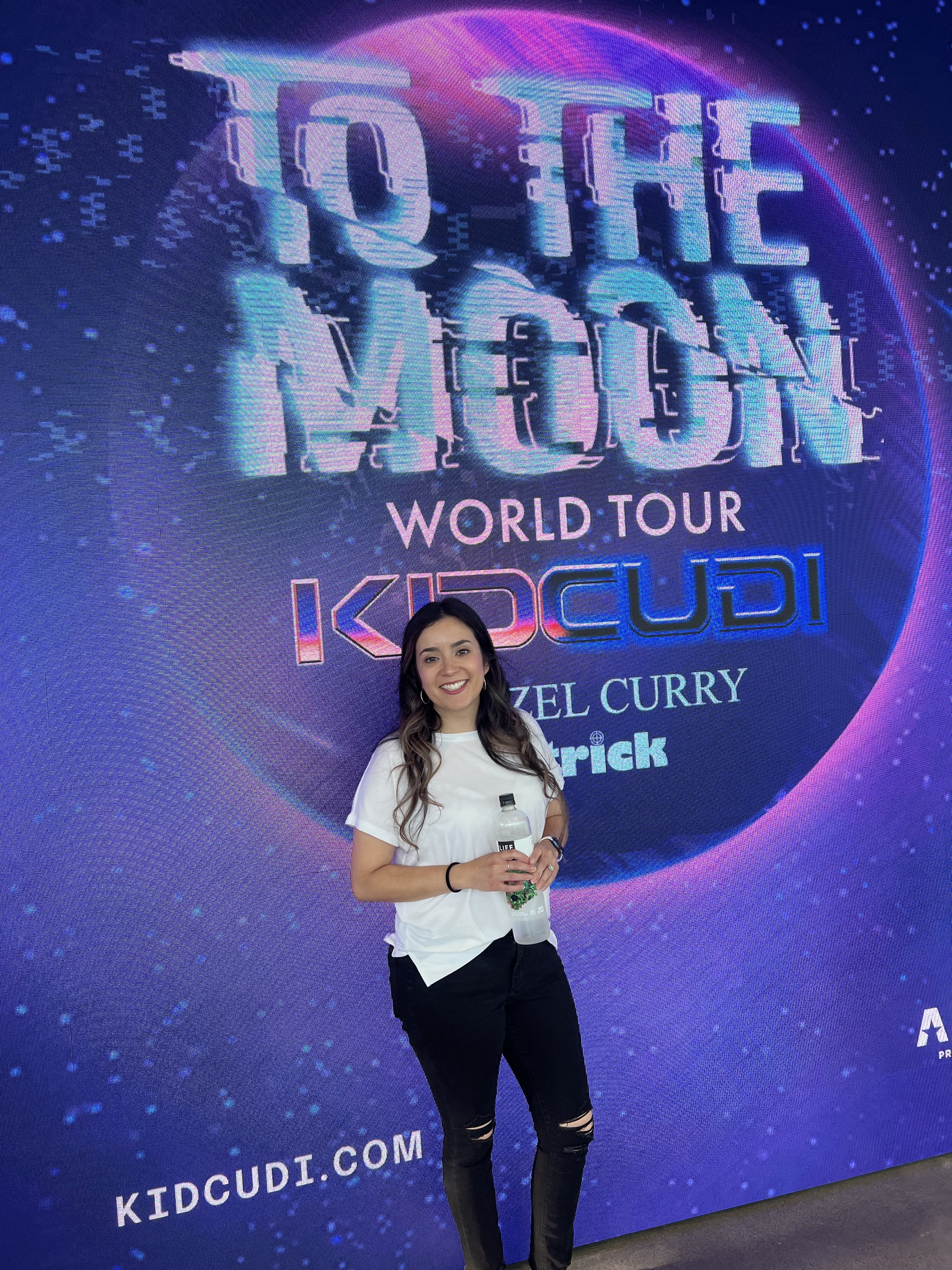 A woman standing in front of a large colorful digital display that reads 'To the Moon' and promotes a world tour for Kid Cudi. She is smiling and holding a water bottle.