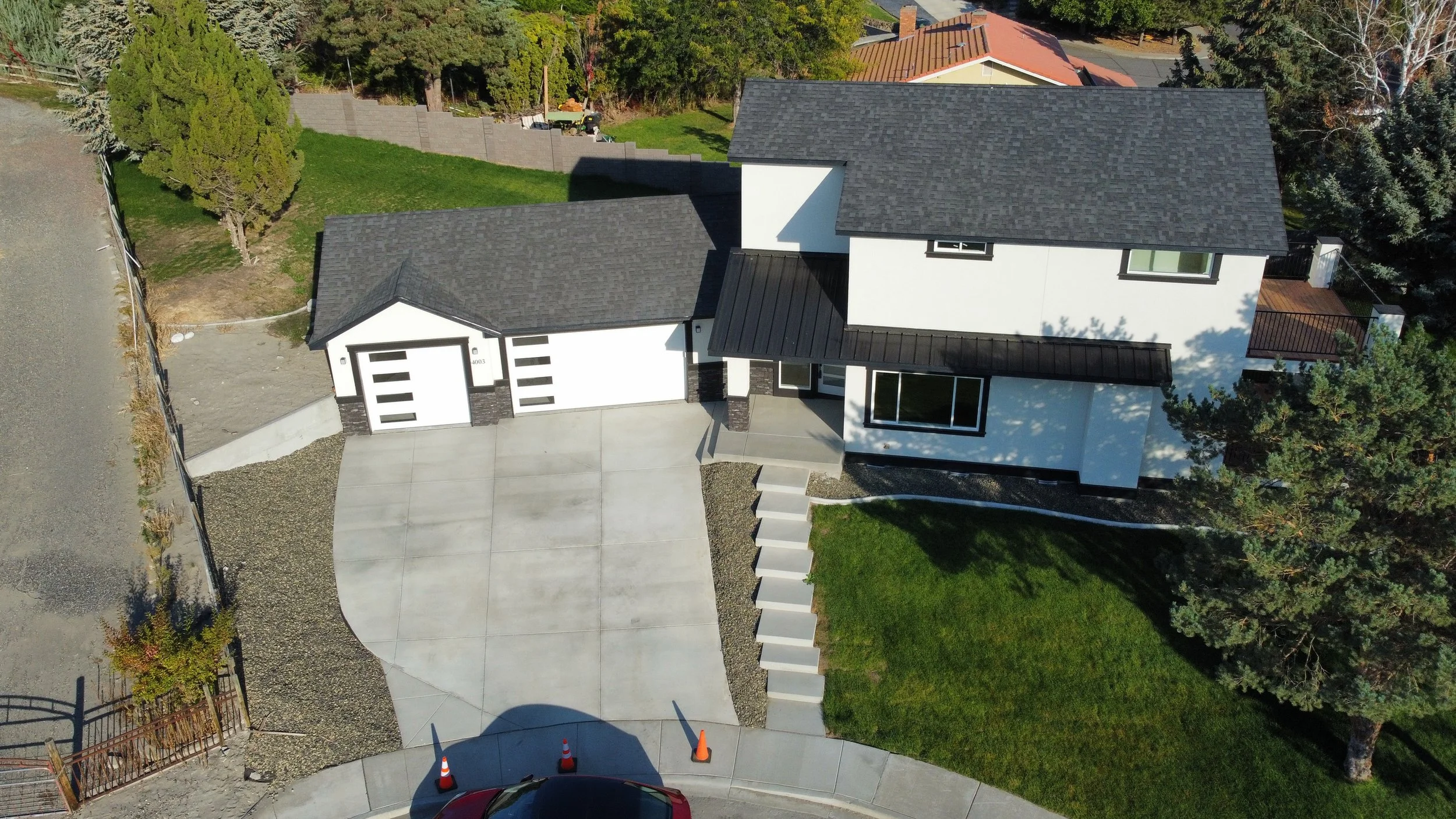 Aerial view of a modern white house with black roofing, a concrete driveway, and a front lawn. There are trees and neighboring houses in the background.