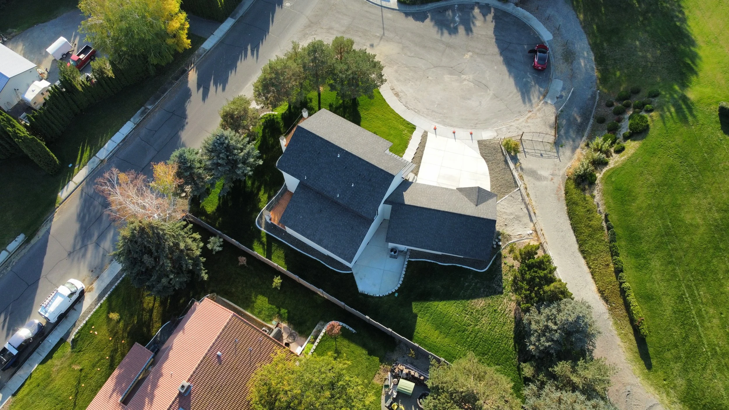 Aerial view of a house with a gray roof surrounded by green lawns, trees, and a fence. There is a circular driveway with a red car parked, and nearby is a gravel pathway. Adjacent properties and streets are visible.