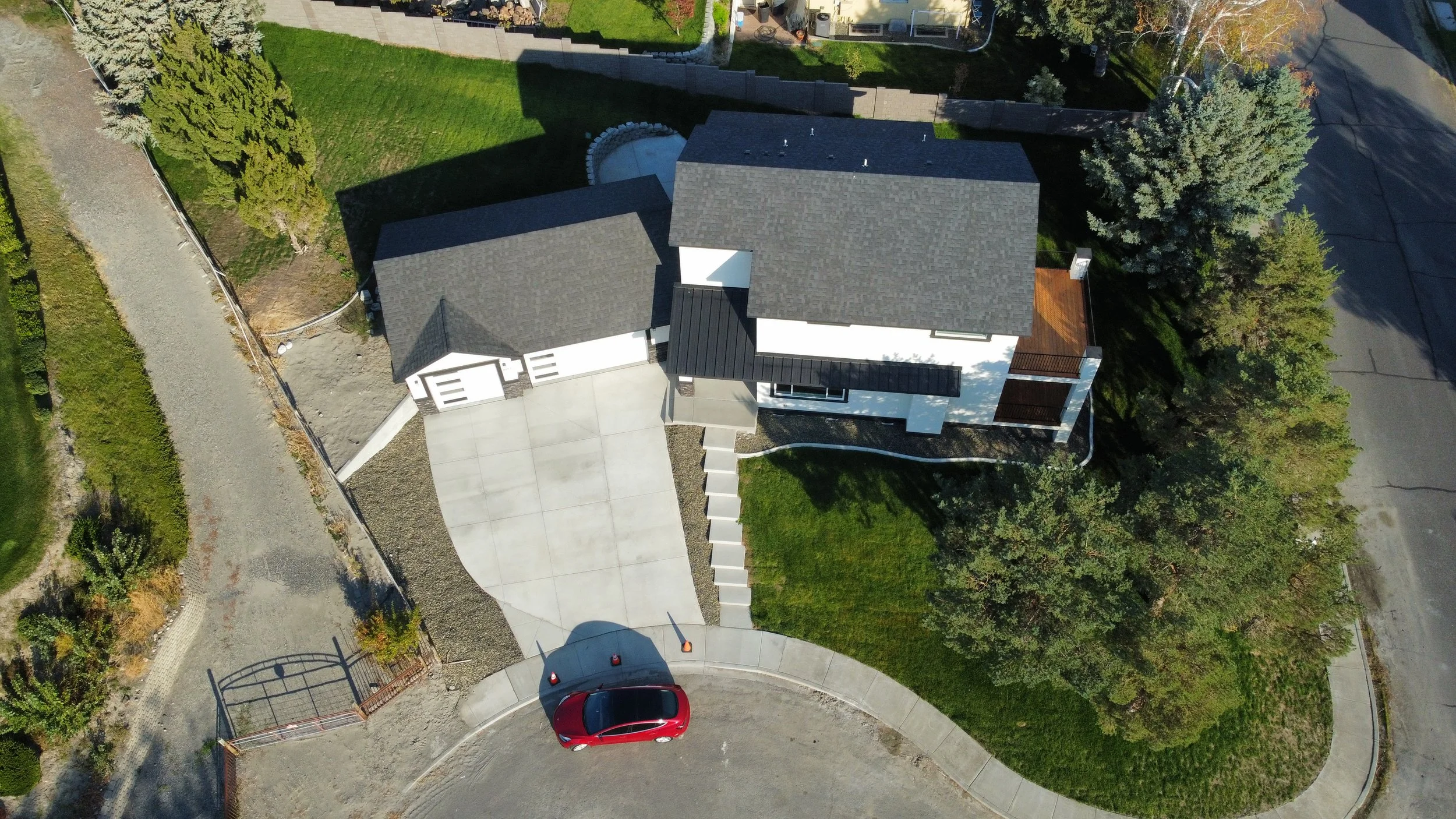 Aerial view of a modern house with a gray roof, white walls, and an attached garage, surrounded by trees, a grassy yard, and a concrete driveway with a red car parked on it.