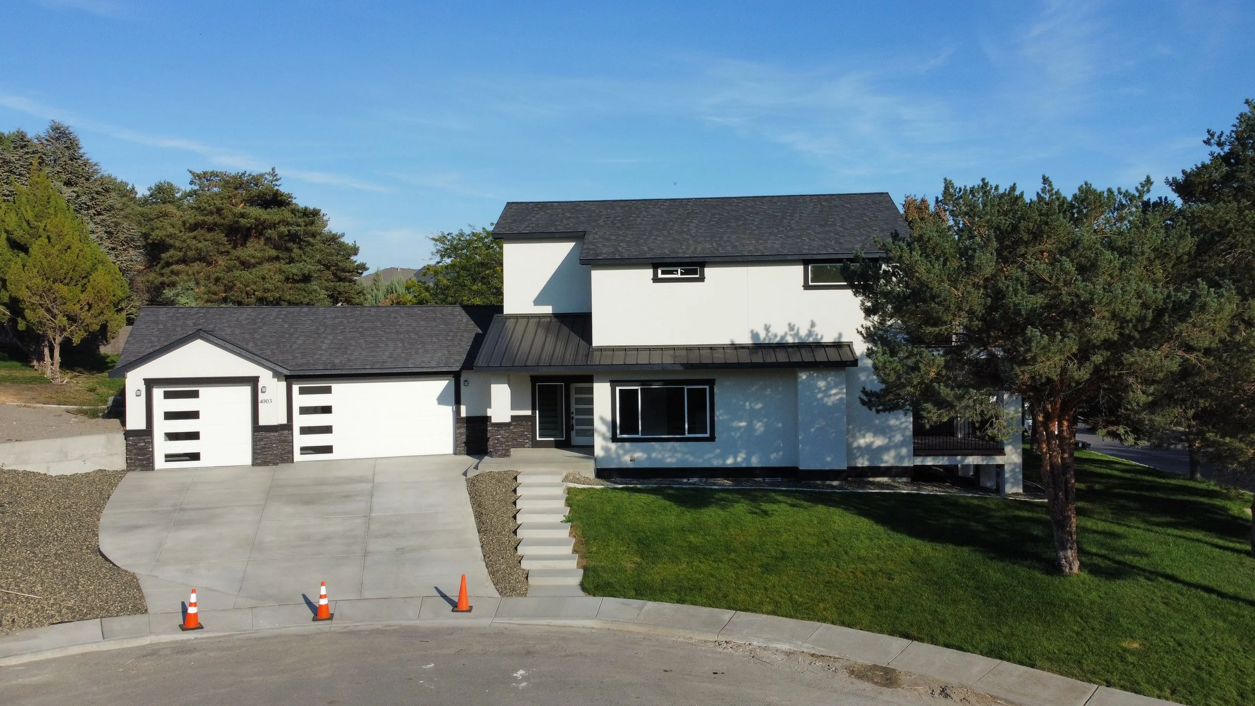 Modern two-story house with white exterior, black roof, and attached garage, surrounded by trees and a well-maintained lawn, with orange construction cones on the driveway.