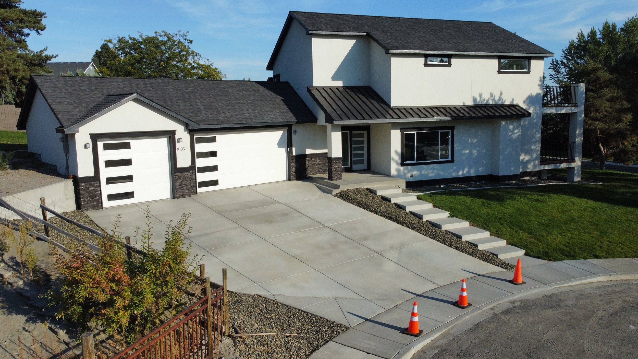 Modern two-story house with a white exterior and black trim, three-car garage, concrete driveway, small front steps, and a well-maintained lawn, with orange traffic cones at the curb.