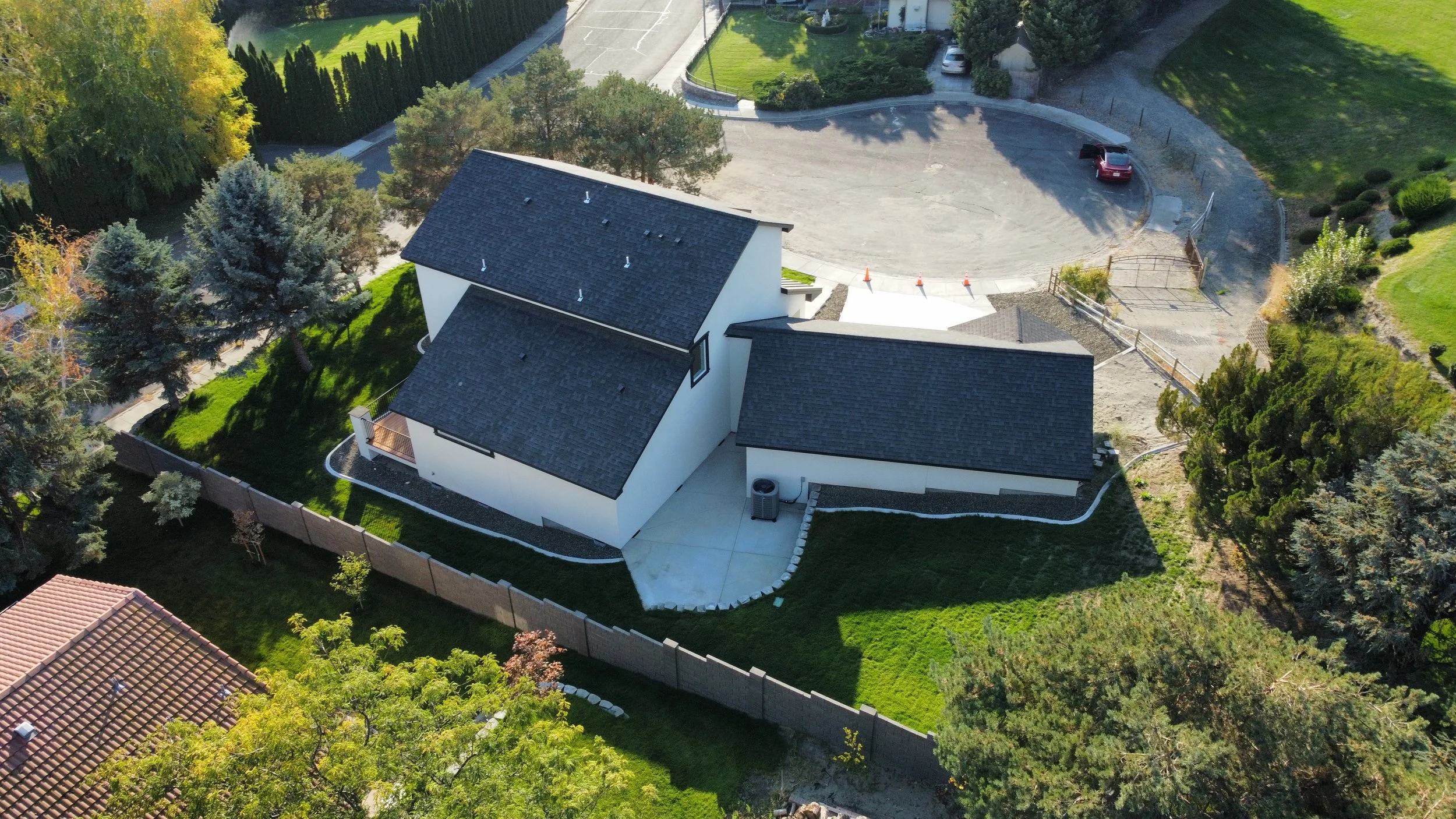 An aerial view of a residential house with a black shingle roof, white exterior walls, surrounded by a green lawn, trees, and a fenced backyard. There is a driveway and a parking area with a few cars in the background.