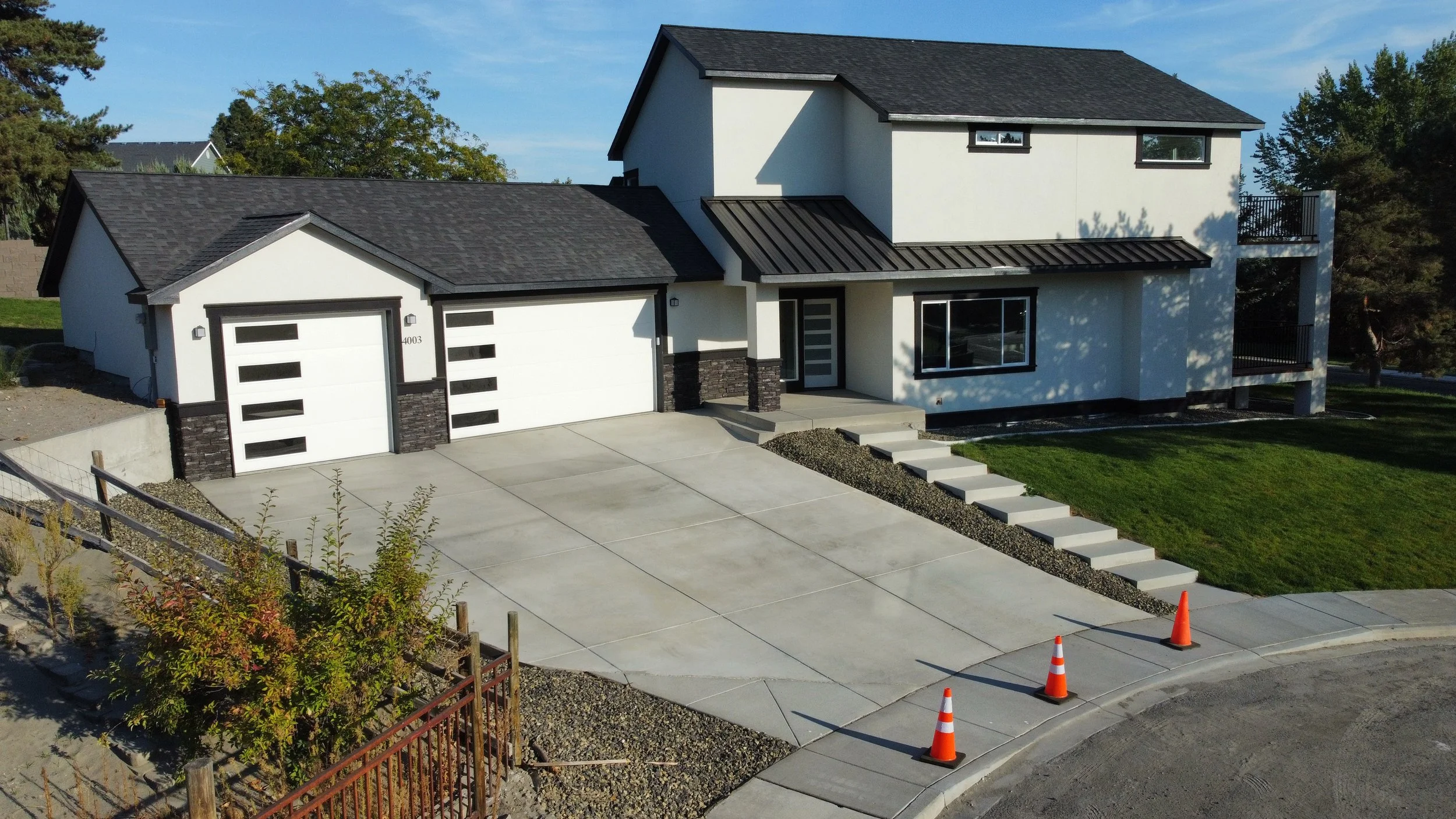 Modern white two-story house with black trim, attached garage with black and white doors, concrete driveway, front steps, and a small porch. Green lawn, trees in background, and orange traffic cones on the sidewalk.
