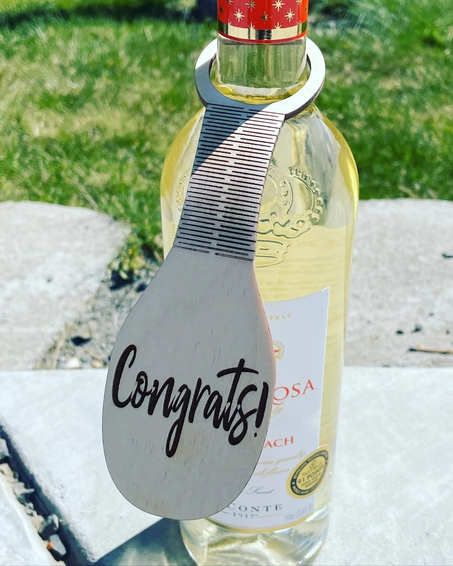 A bottle of white wine with a wooden tag attached that reads 'Congrats!' hanging from the neck of the bottle, outdoors on a gray surface with green grass in the background.