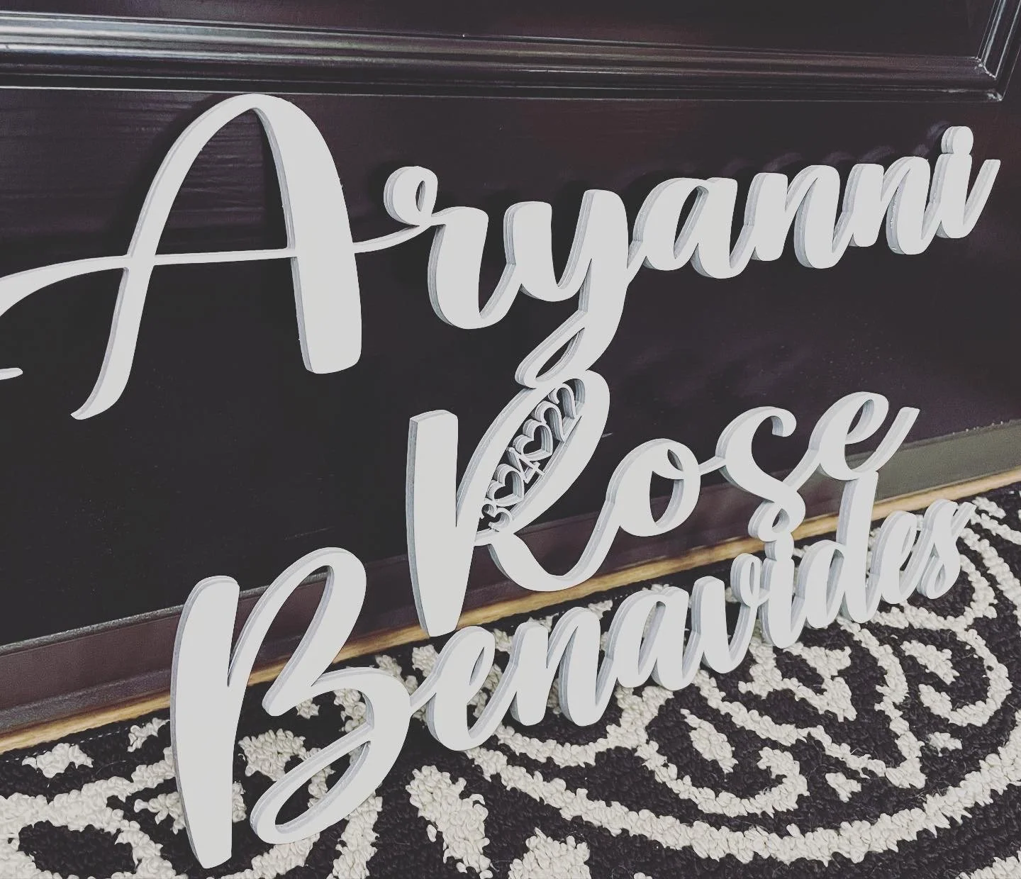 White decorative sign with the words 'A yyamii & Kosei Bemarried' in cursive, leaning against a dark wood background, placed on a black and white patterned rug.