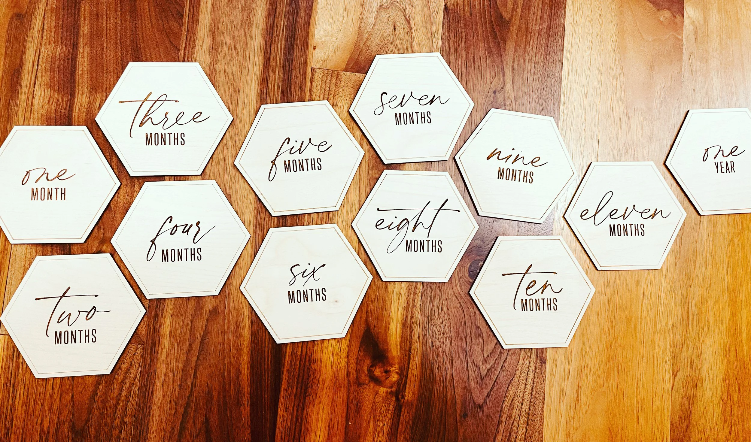 A collection of white hexagon and octagon-shaped cards with handwritten-style text displaying the months and years, arranged on a wooden surface. The cards show months from one month to eleven months, and two cards indicate one year.