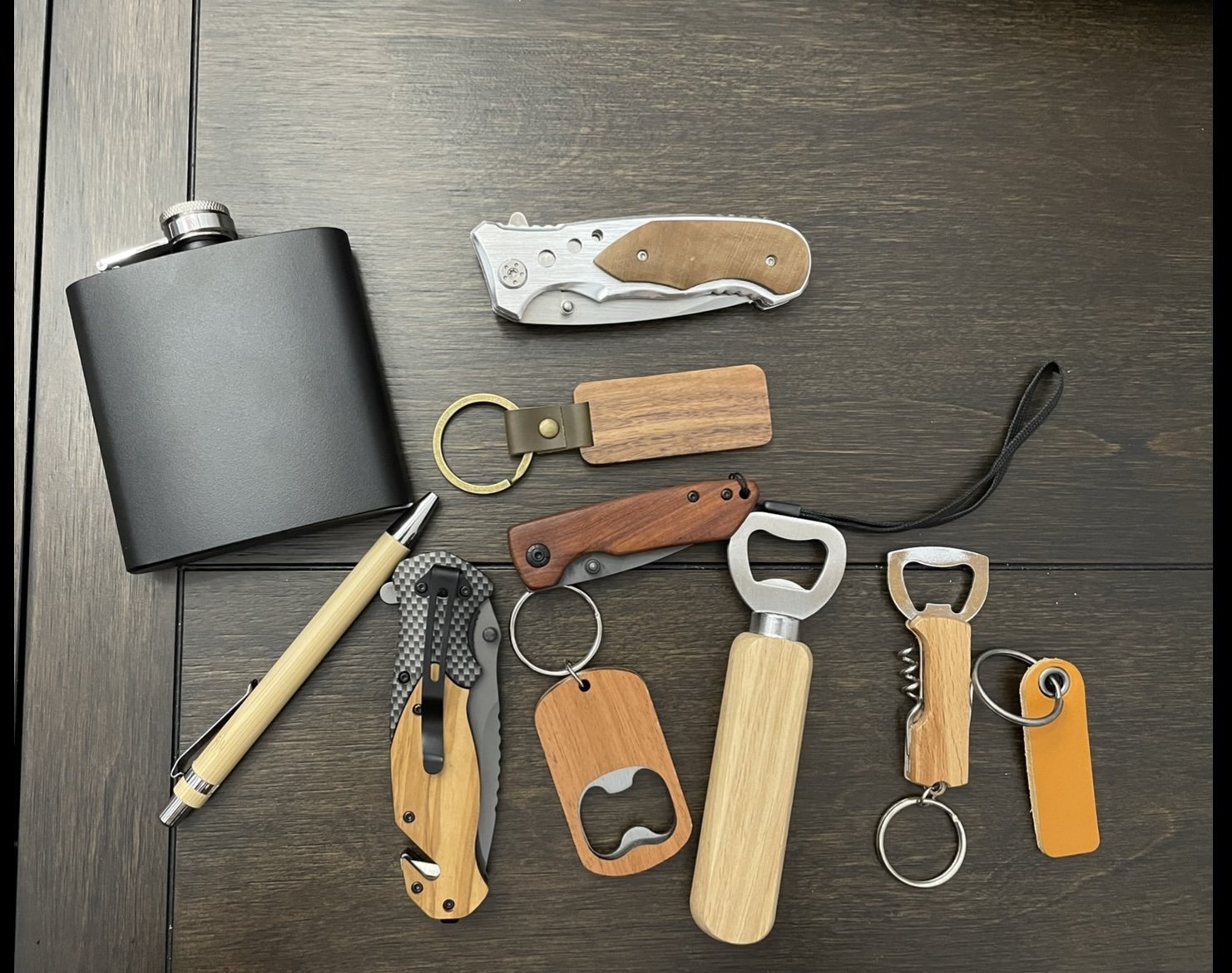 Collection of various pocket knives, keychain tools, and a black flask with a pen on a dark wooden surface.