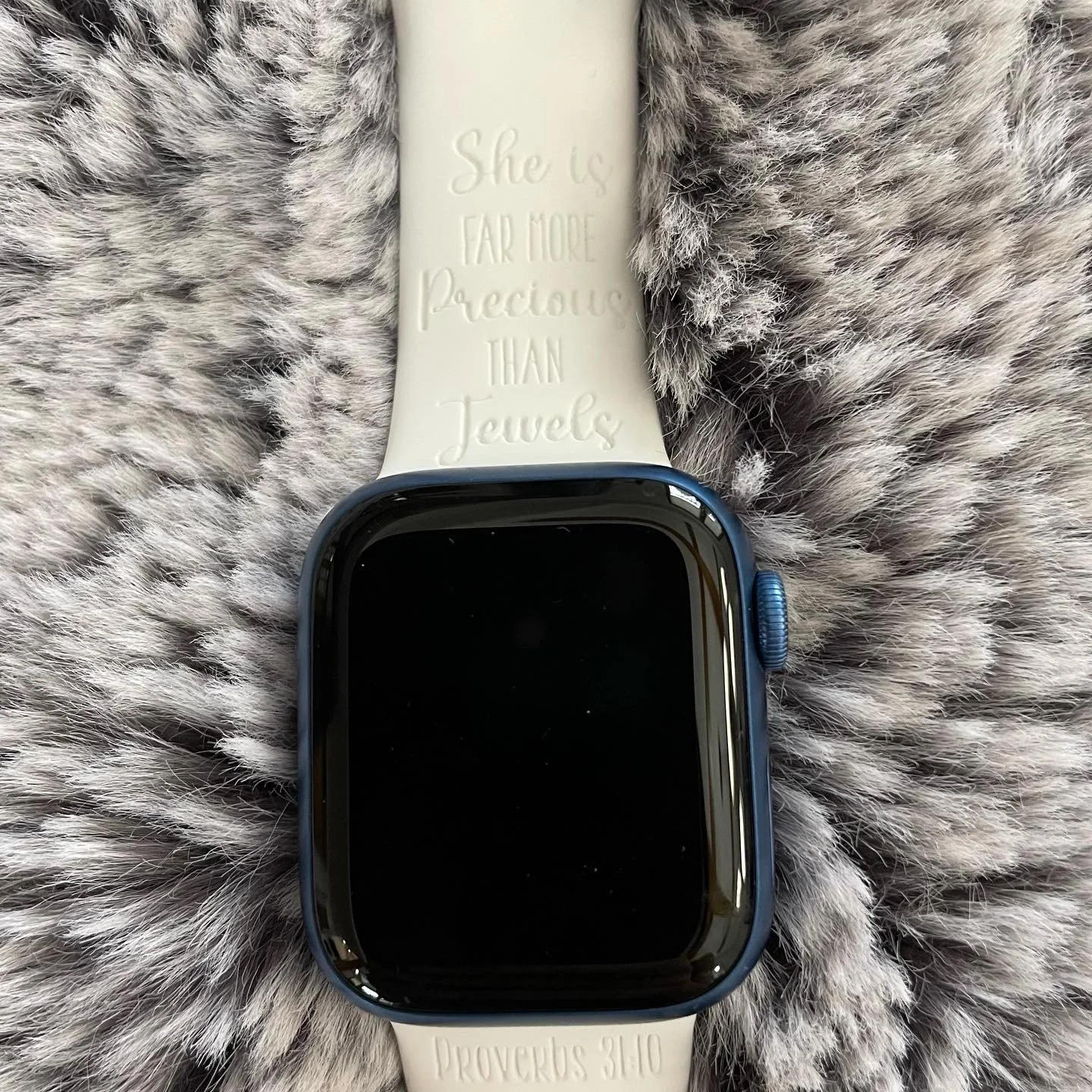 A smartwatch with a black screen and a blue case, placed on a fluffy gray and white faux fur surface. The smartwatch has a light-colored band with embossed text that reads, 'She is FAR MORE Precious THAN Jewels' and 'Proverbs 31:10'.