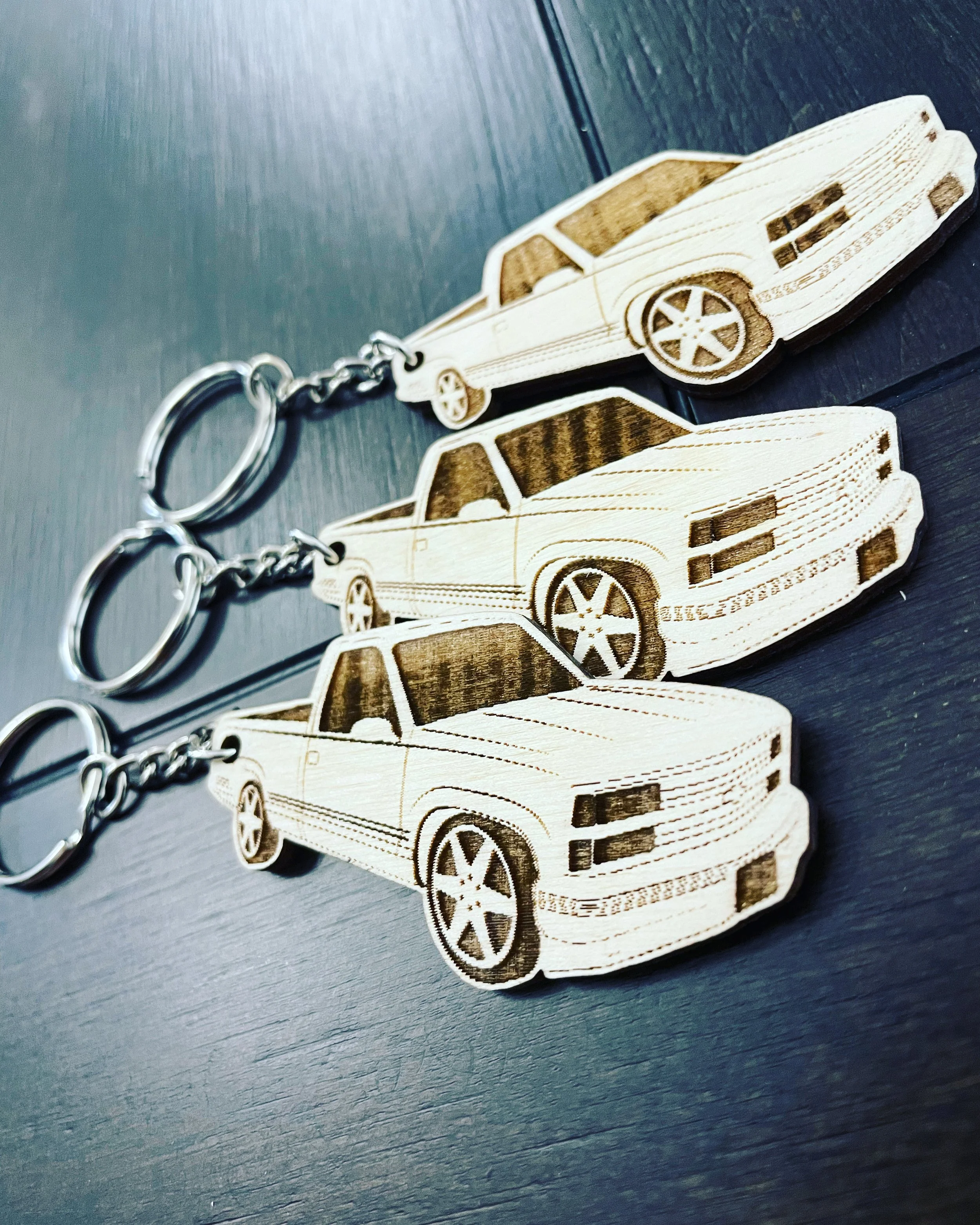 Three wooden car keychains with a car design, attached to metal keyrings, lying on a dark surface.