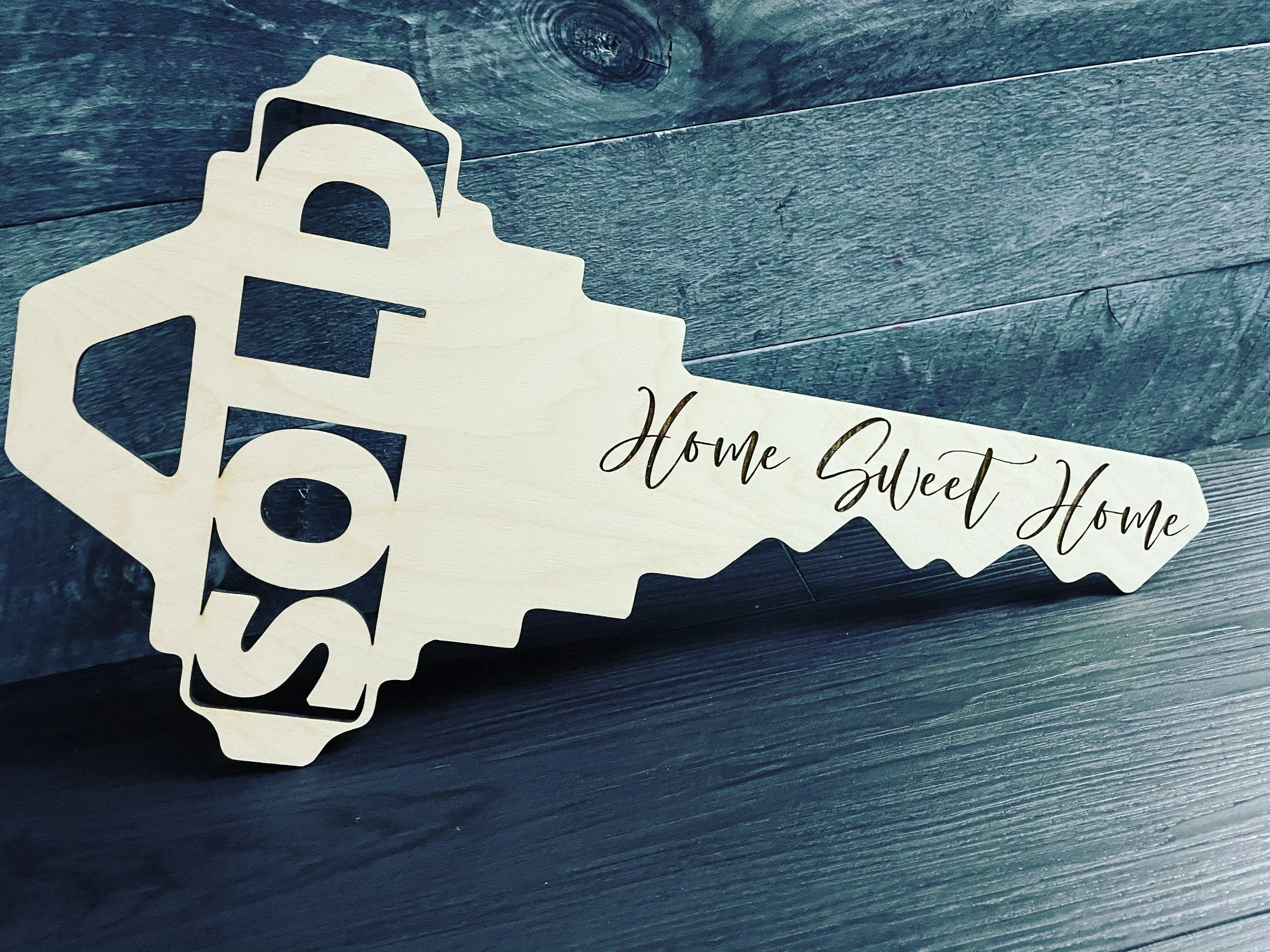 Decorative wooden key with the word "HOME" cut out, attached to a wooden background, with the phrase "Home Sweet Home" written on it.