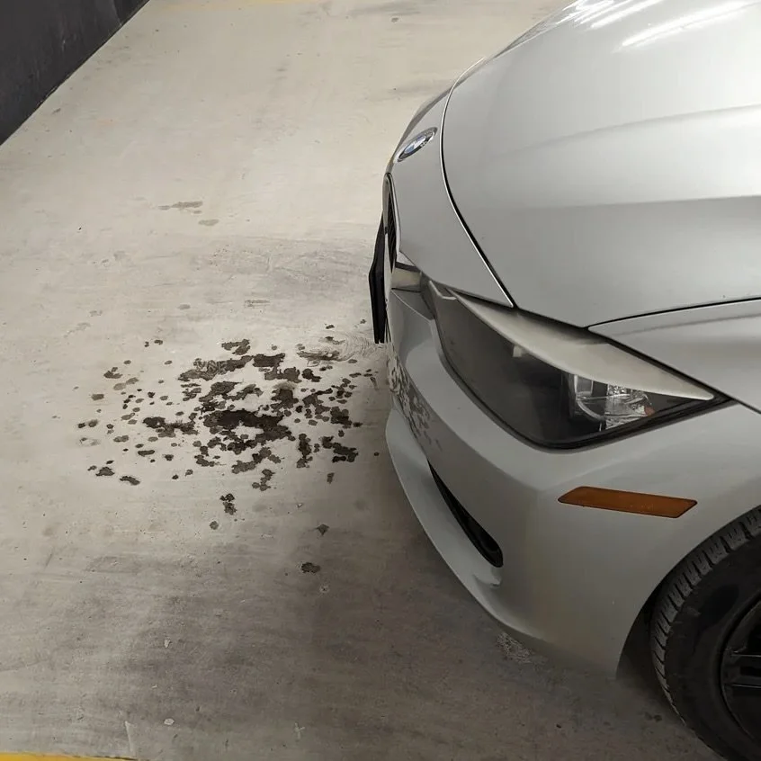 A silver BMW parked in a garage with oil spots on the concrete floor below the car