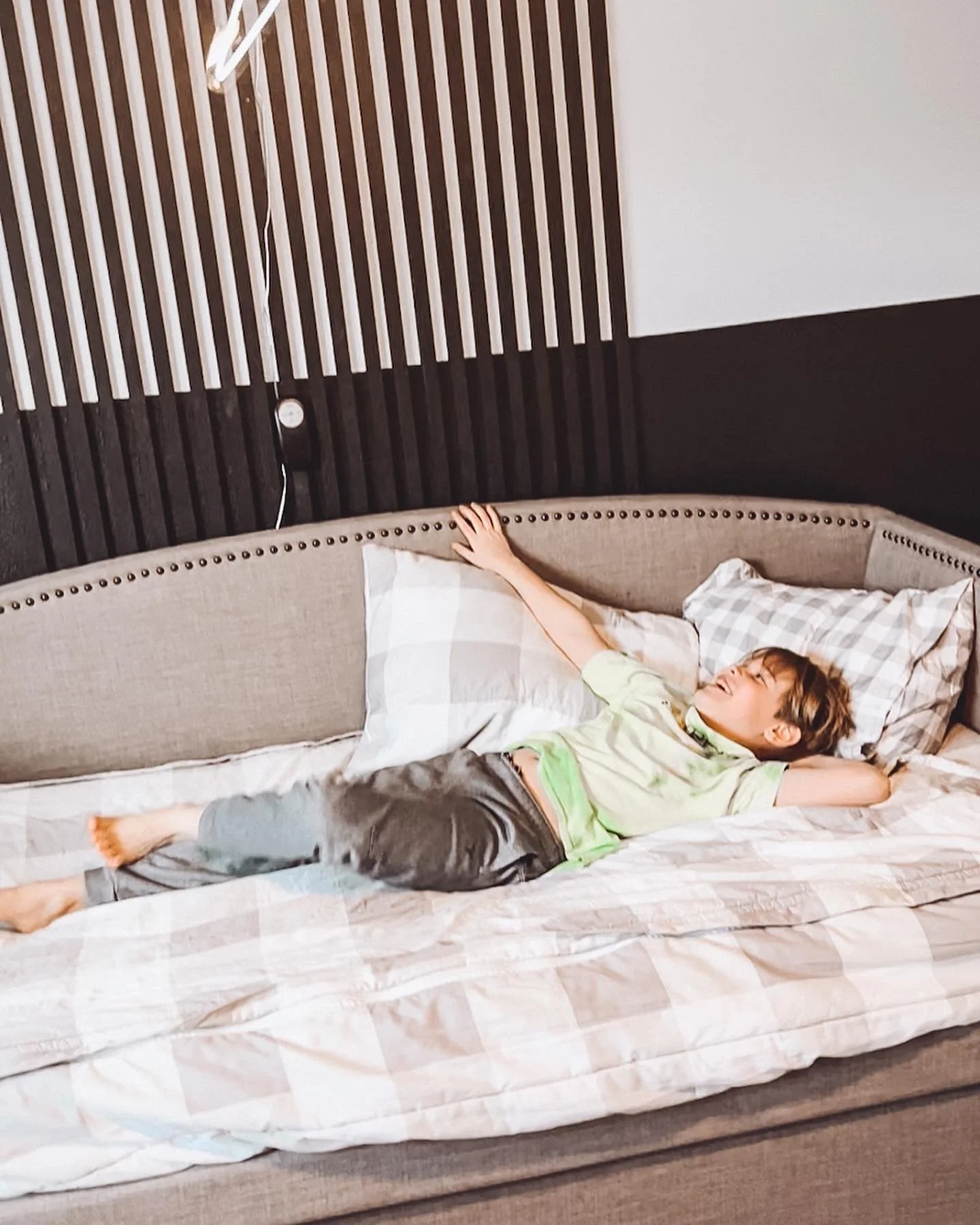A child reclining on a bed in a bedroom with a wood slat accent wall and a wall-mounted reading lamp.