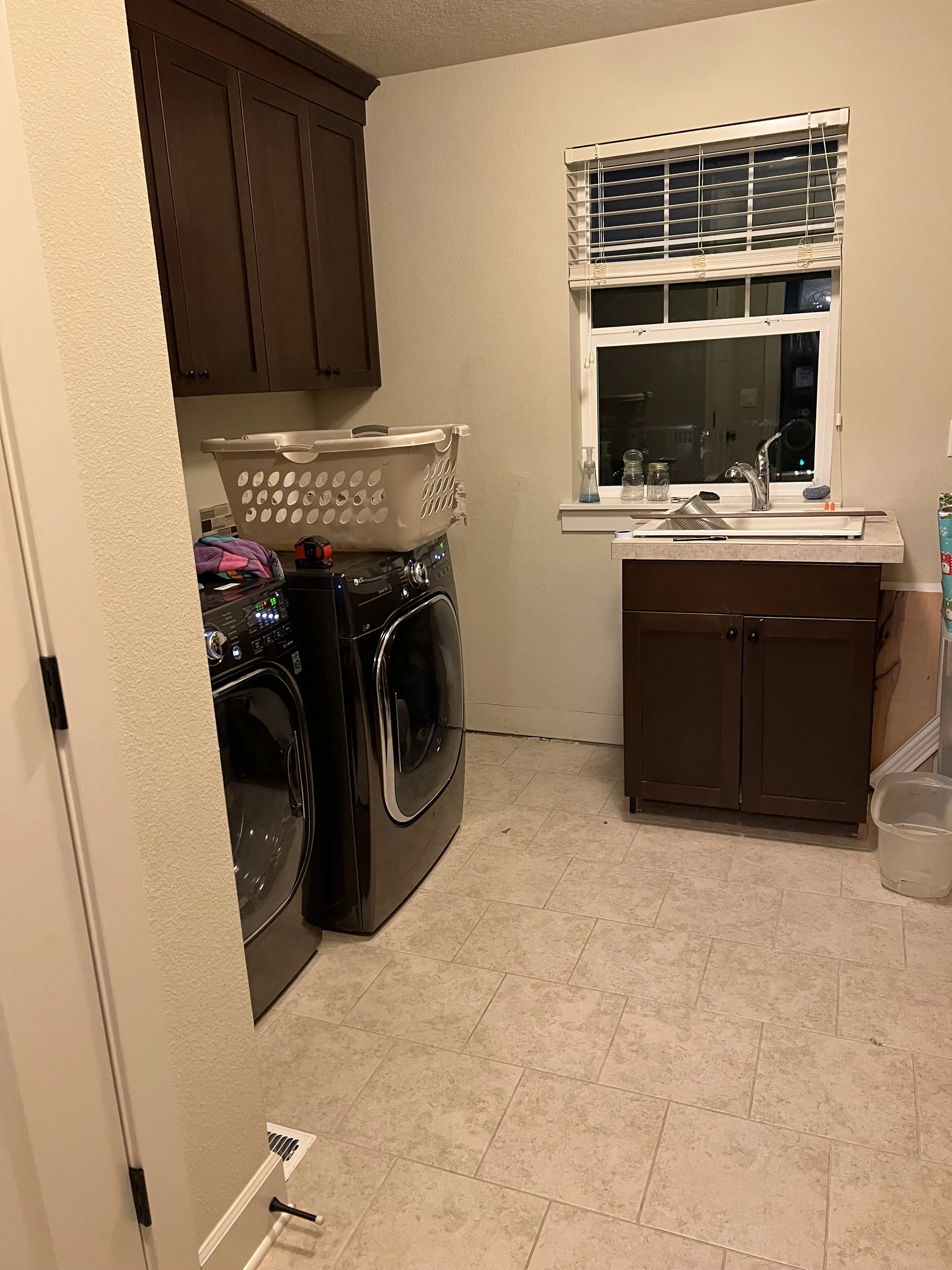 Before renovation: a dated laundry room with brown cabinets, a small utility sink, and builder-grade tile flooring