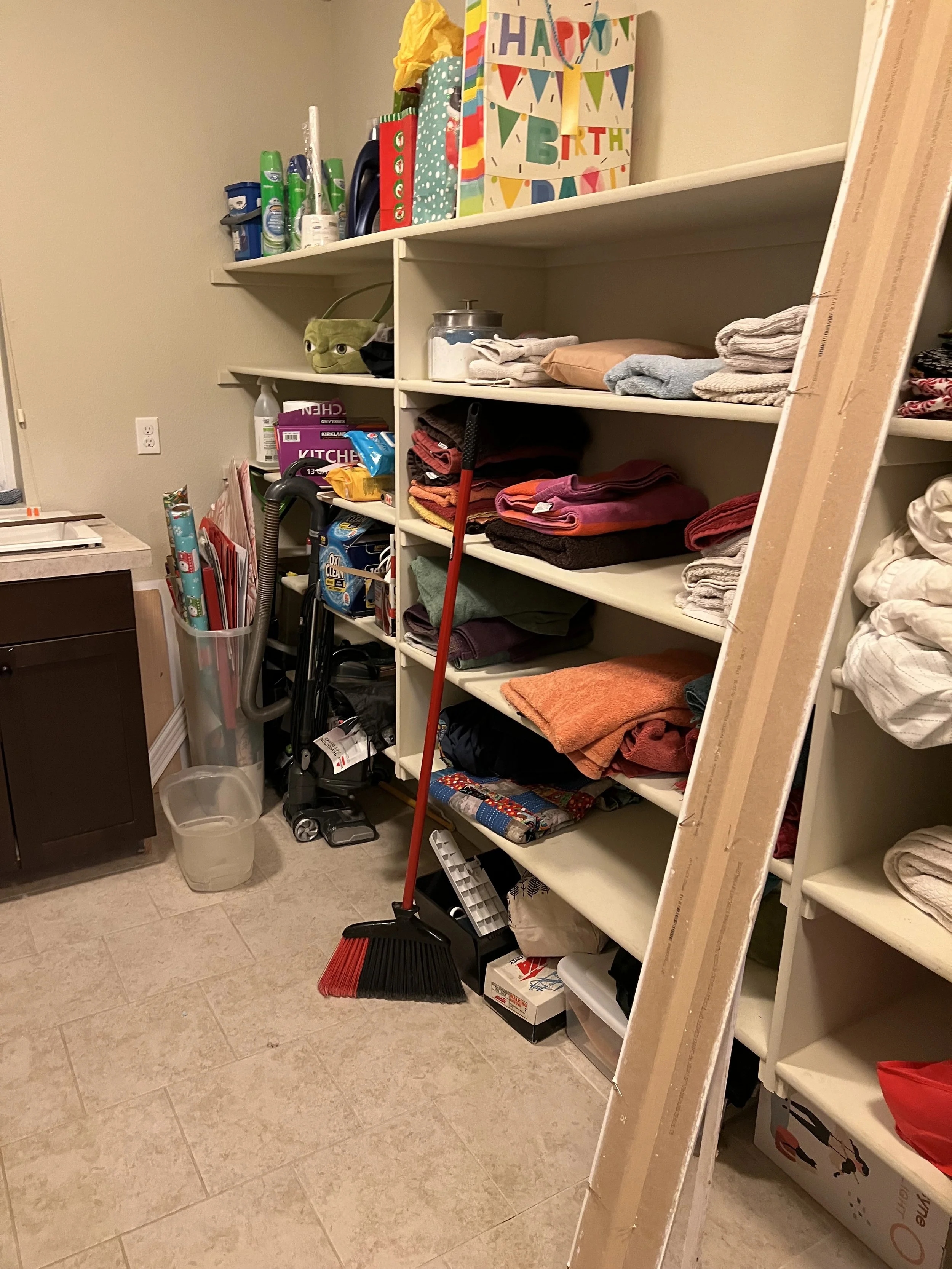 Before renovation: a laundry room with open wood shelving crowded with linens and supplies.