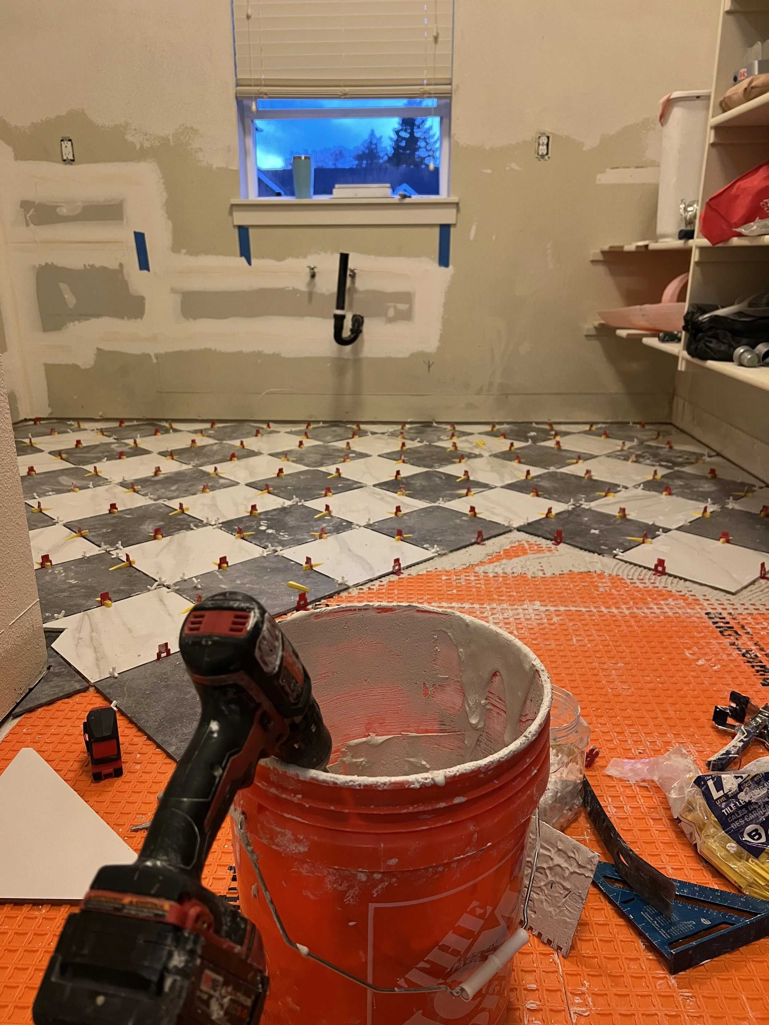 Mid renovation: Grey and white checkerboard tile being laid on the laundry room floor with spacers and tools in frame.