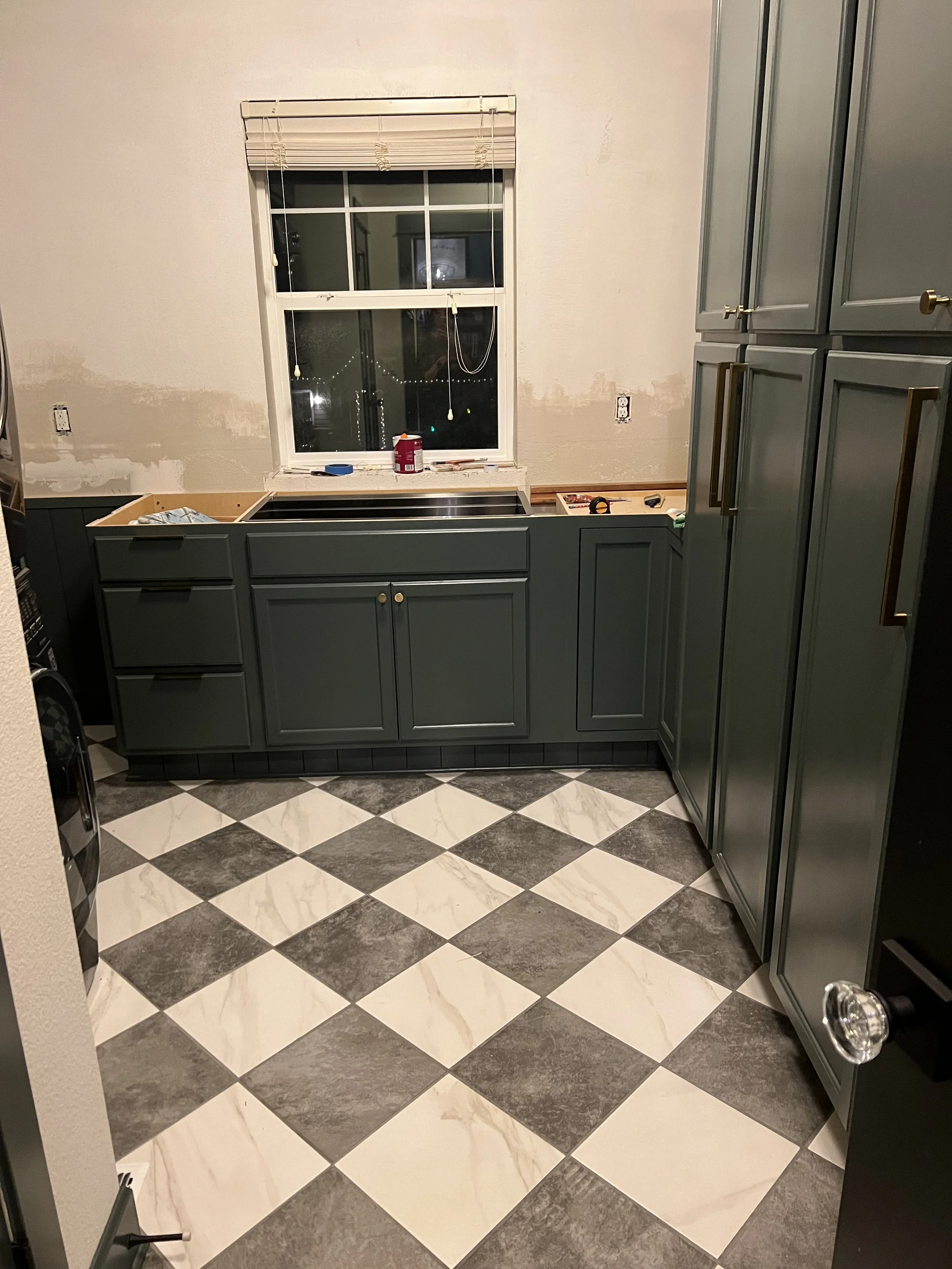 Mid renovation: Pewter green cabinets and brass hardware installed along the laundry room wall above the new checkerboard floor