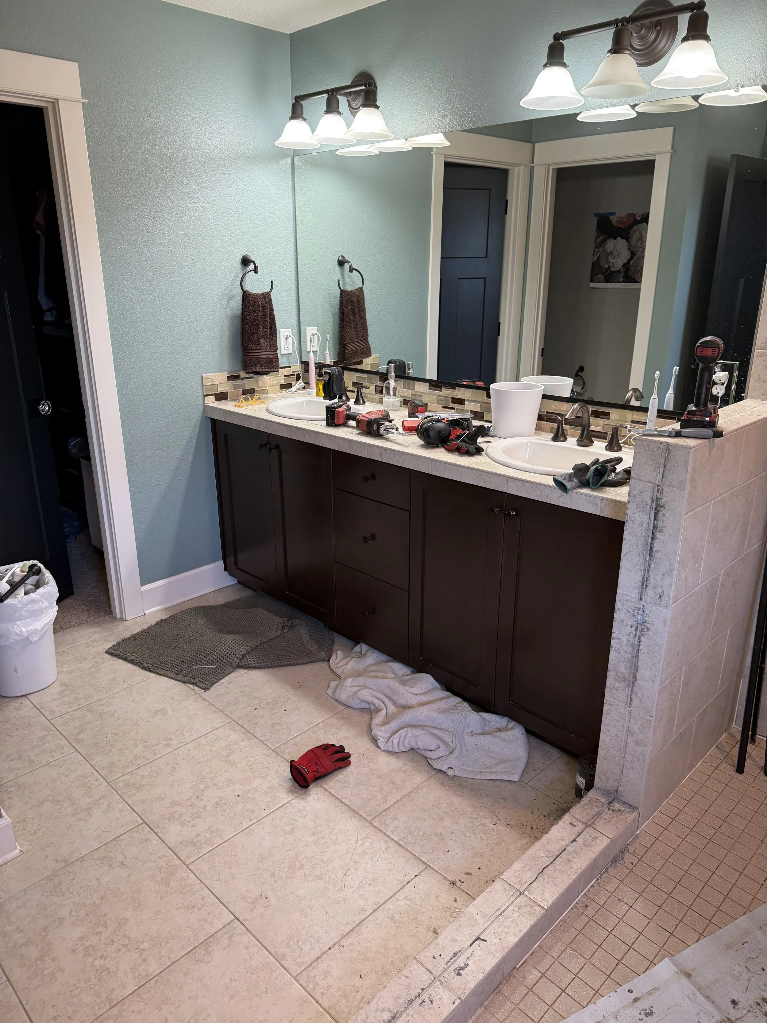 Before renovation: a master bathroom with a dark brown double vanity, a large mirror, teal walls, and a tile floor.
