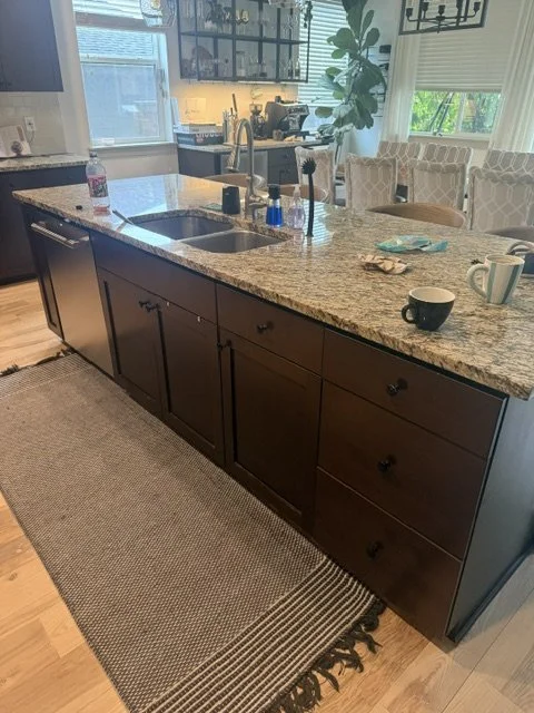 Before renovation: a kitchen with dark brown cabinets, granite countertops, and a long island with a double sink.