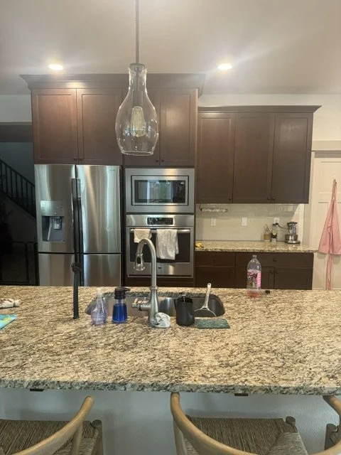 Before renovation: a kitchen with dark brown cabinets, granite counters, and a refrigerator beside a staircase
