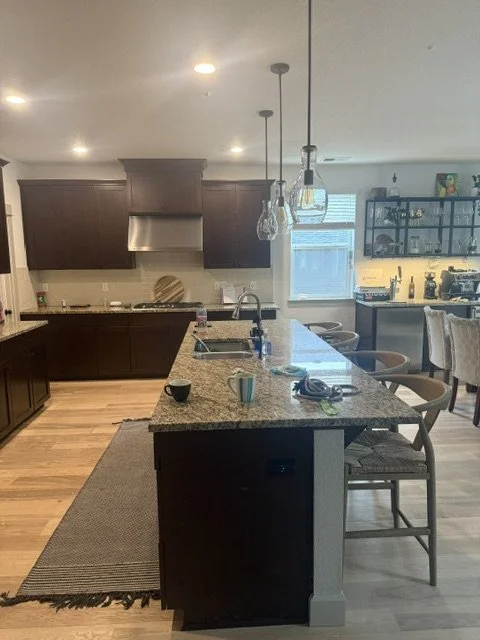 Before renovation: a kitchen bar area with dark brown cabinets, a live-edge wood table, and open shelving with glassware.