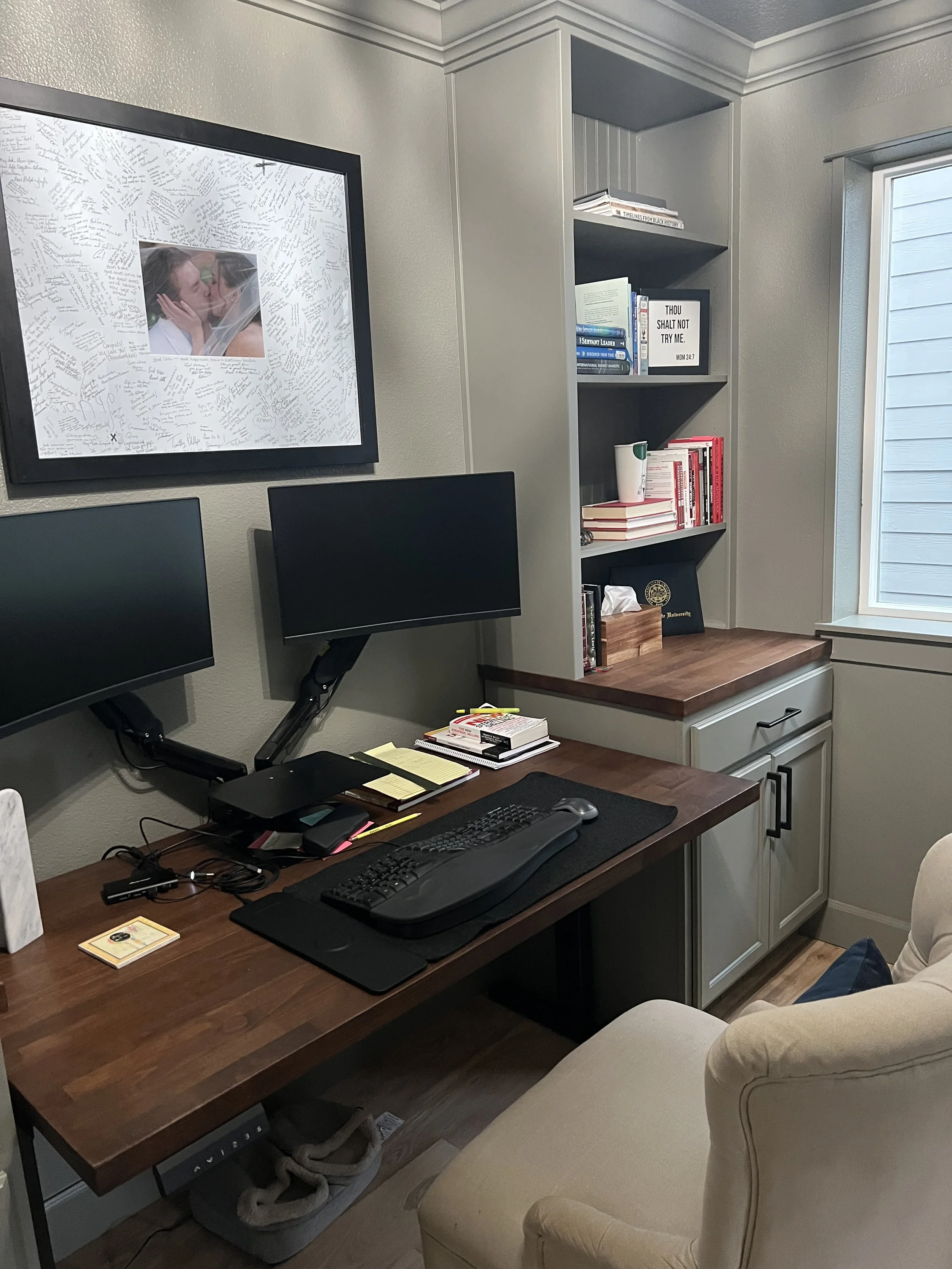 A home office with a custom wood desk, dual monitors, built-in shelving, and a window.