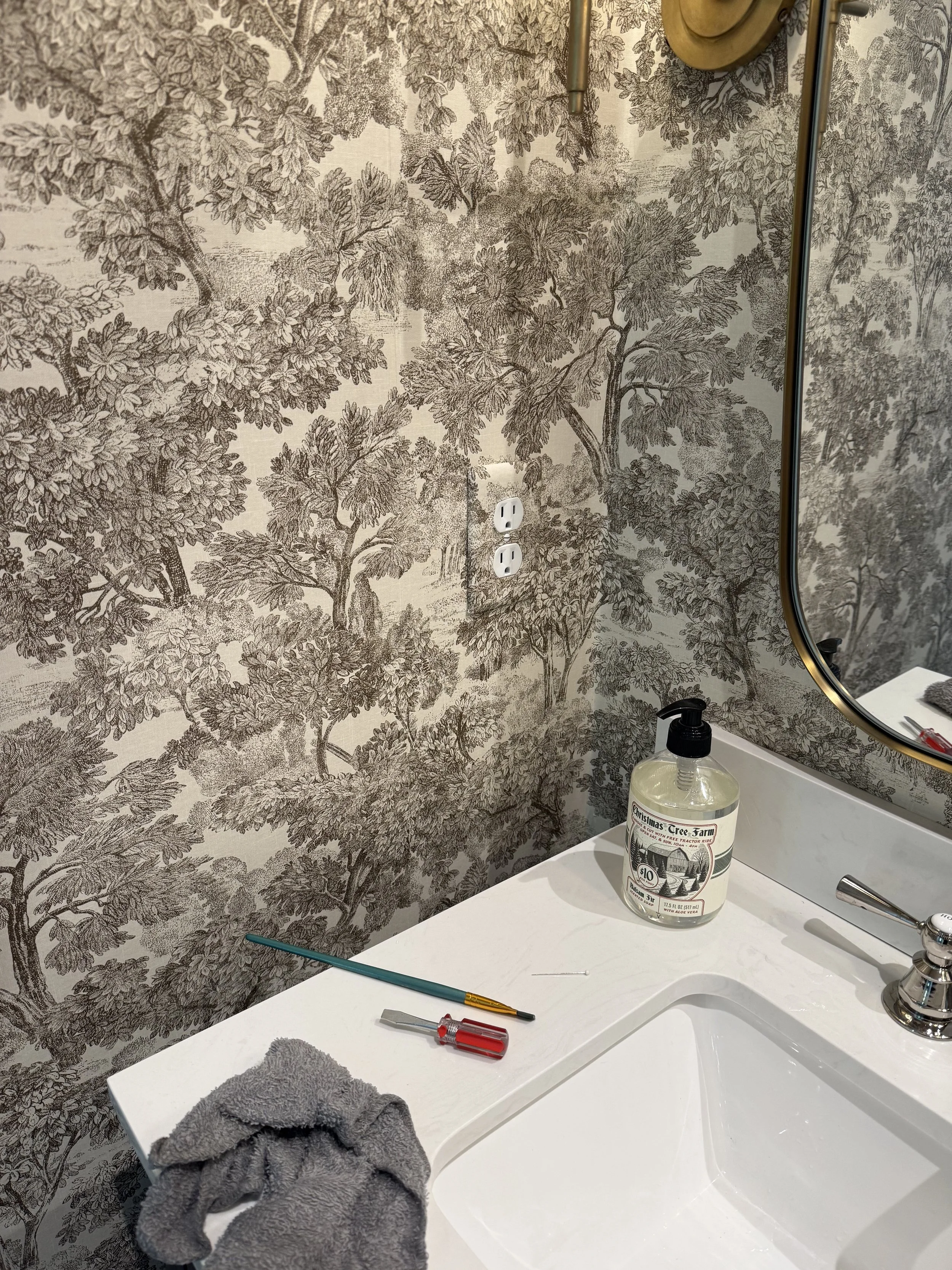 A close view of brown and white botanical wallpaper meeting a white vanity, with install tools on the counter
