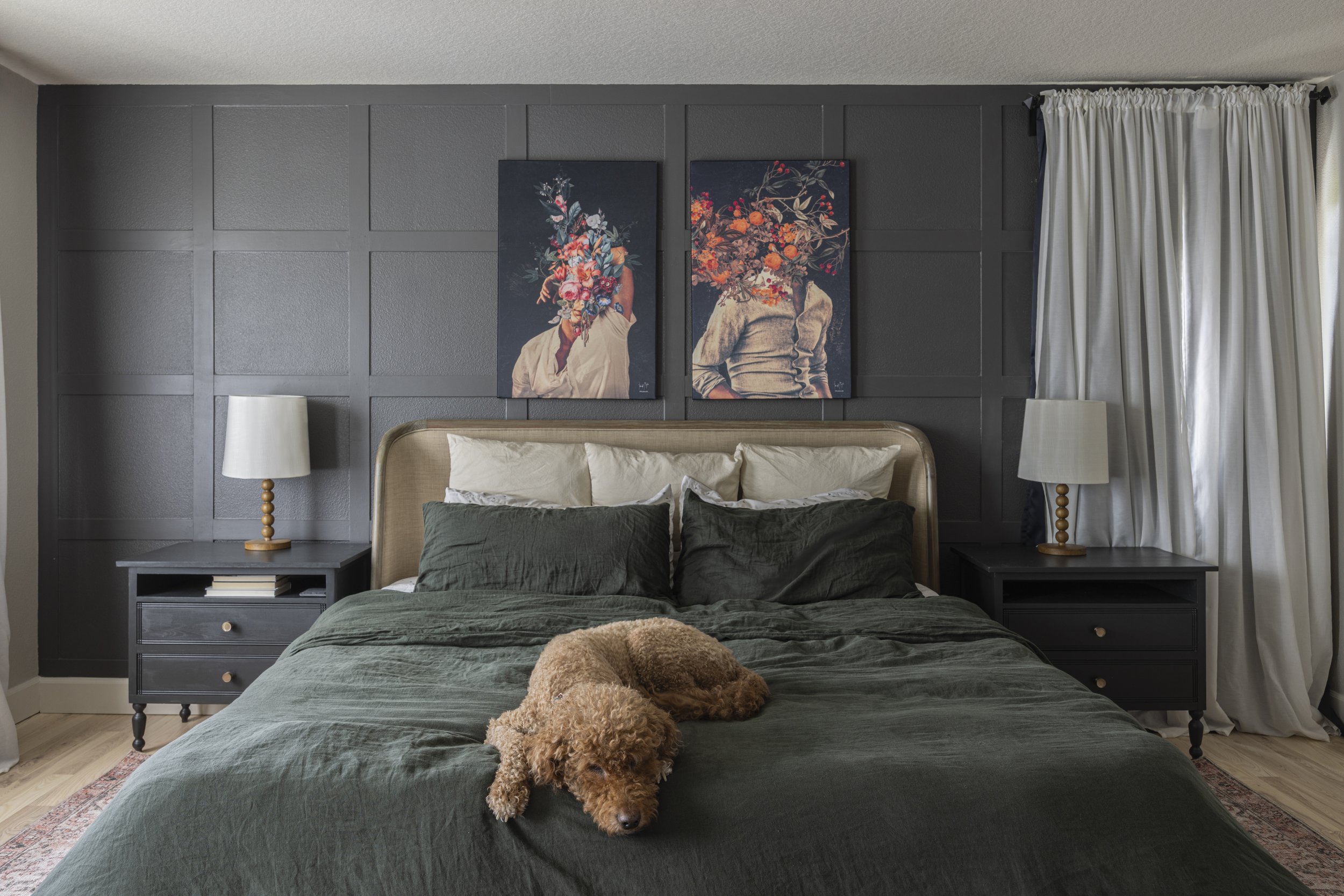 A bedroom with dark green bedding and framed prints above the bed, a labradoodle resting on the duvet, and a curtained window.
