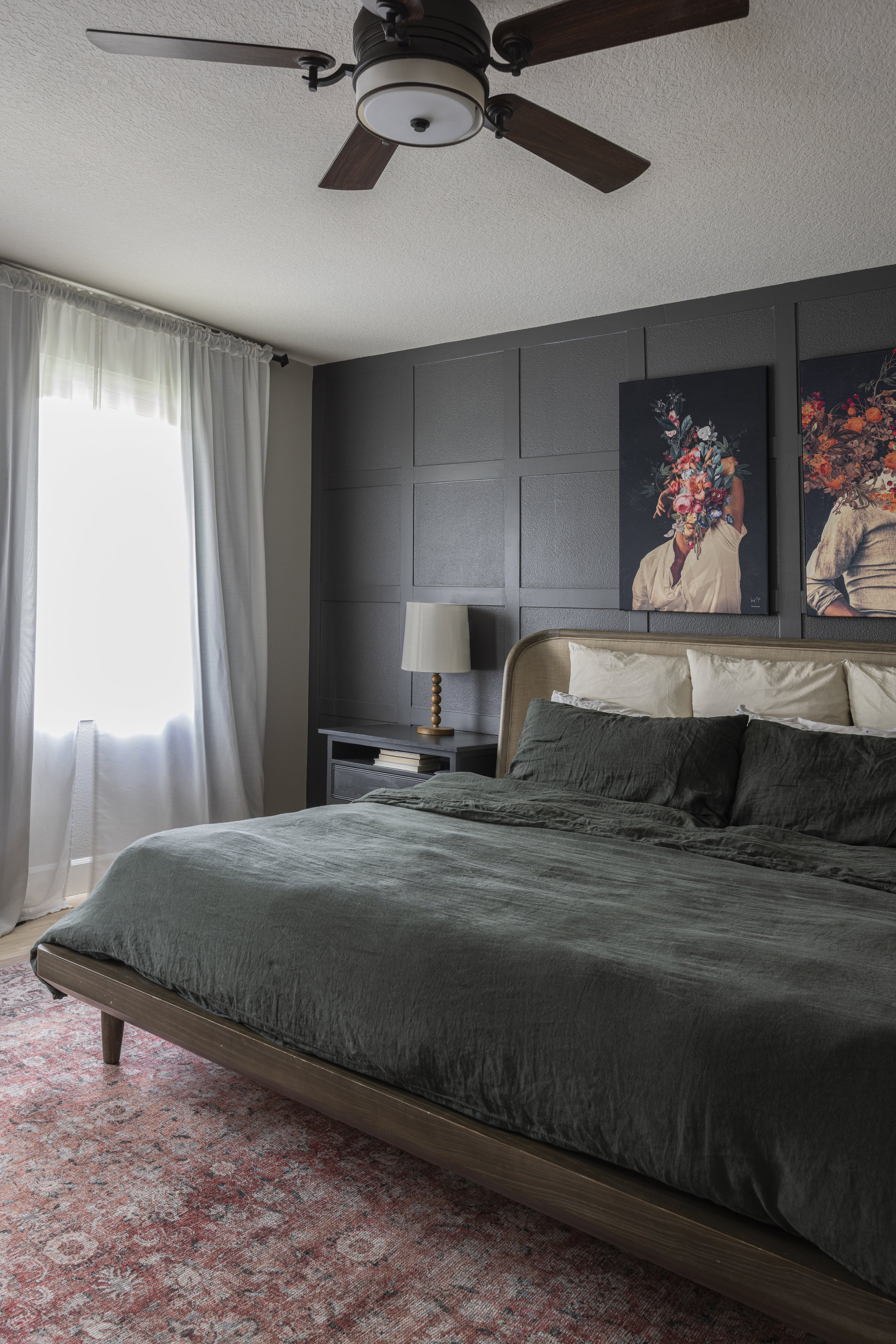 A bedroom with a dark ceiling fan, dark green bedding, and a tall curtained window with a patterned rug.