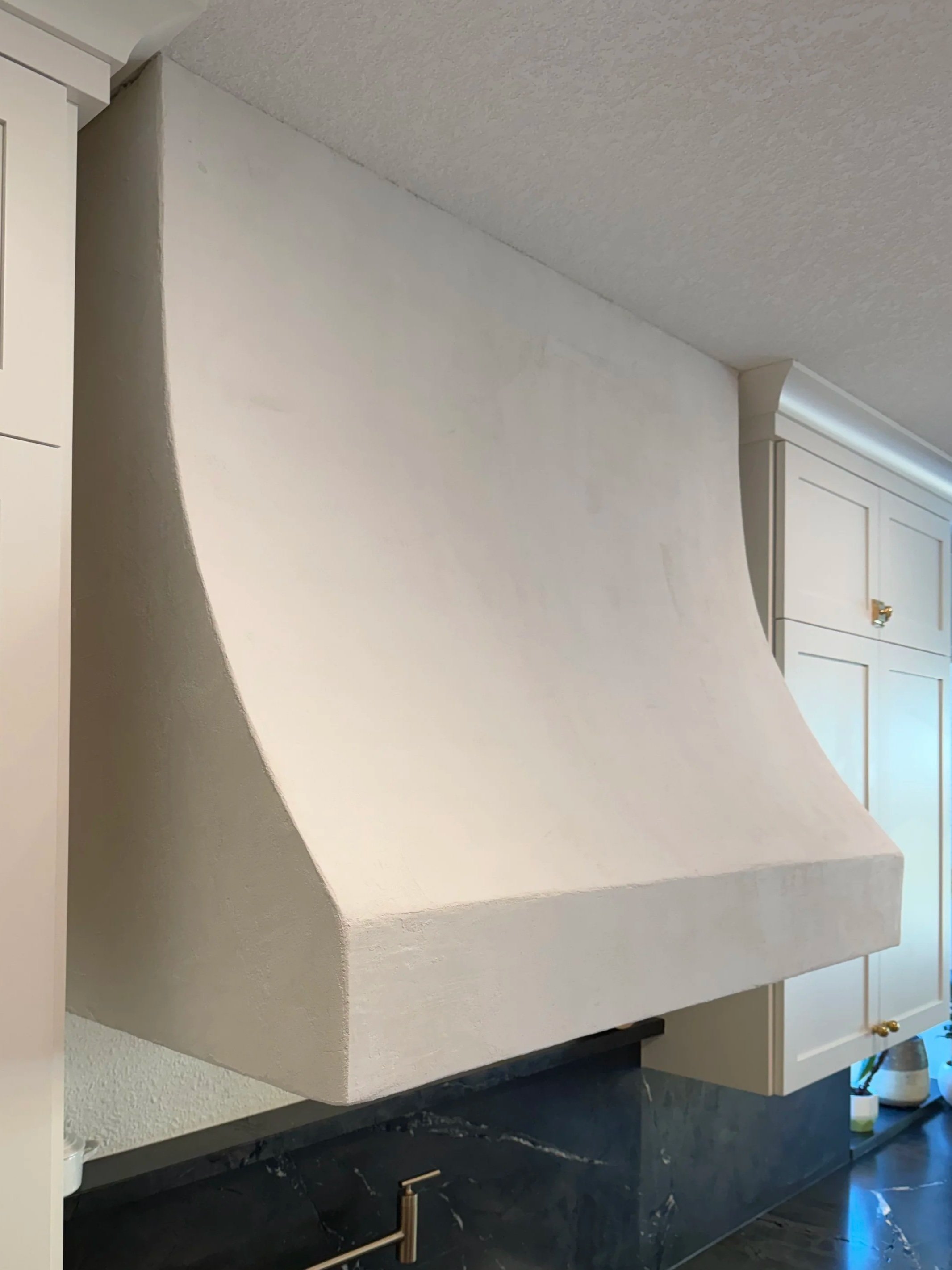 A curved white plaster range hood above a dark countertop, with white cabinetry alongside.