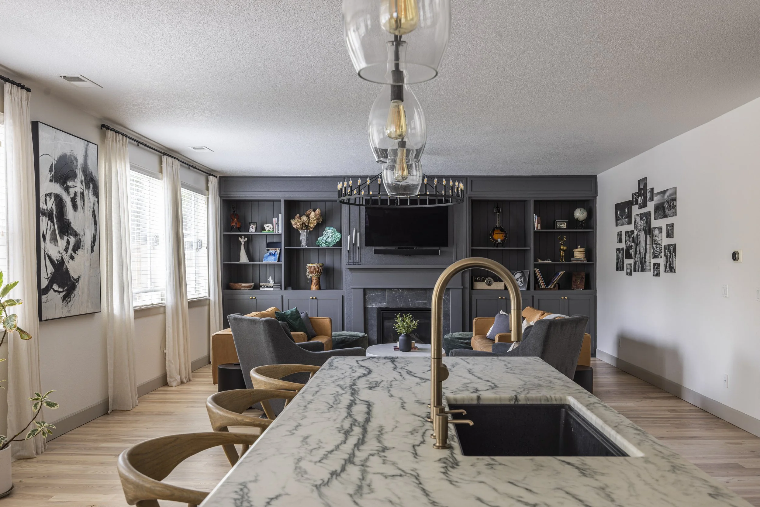A kitchen with a marble countertop island and brass faucet, looking through to a living room with dark built-ins.