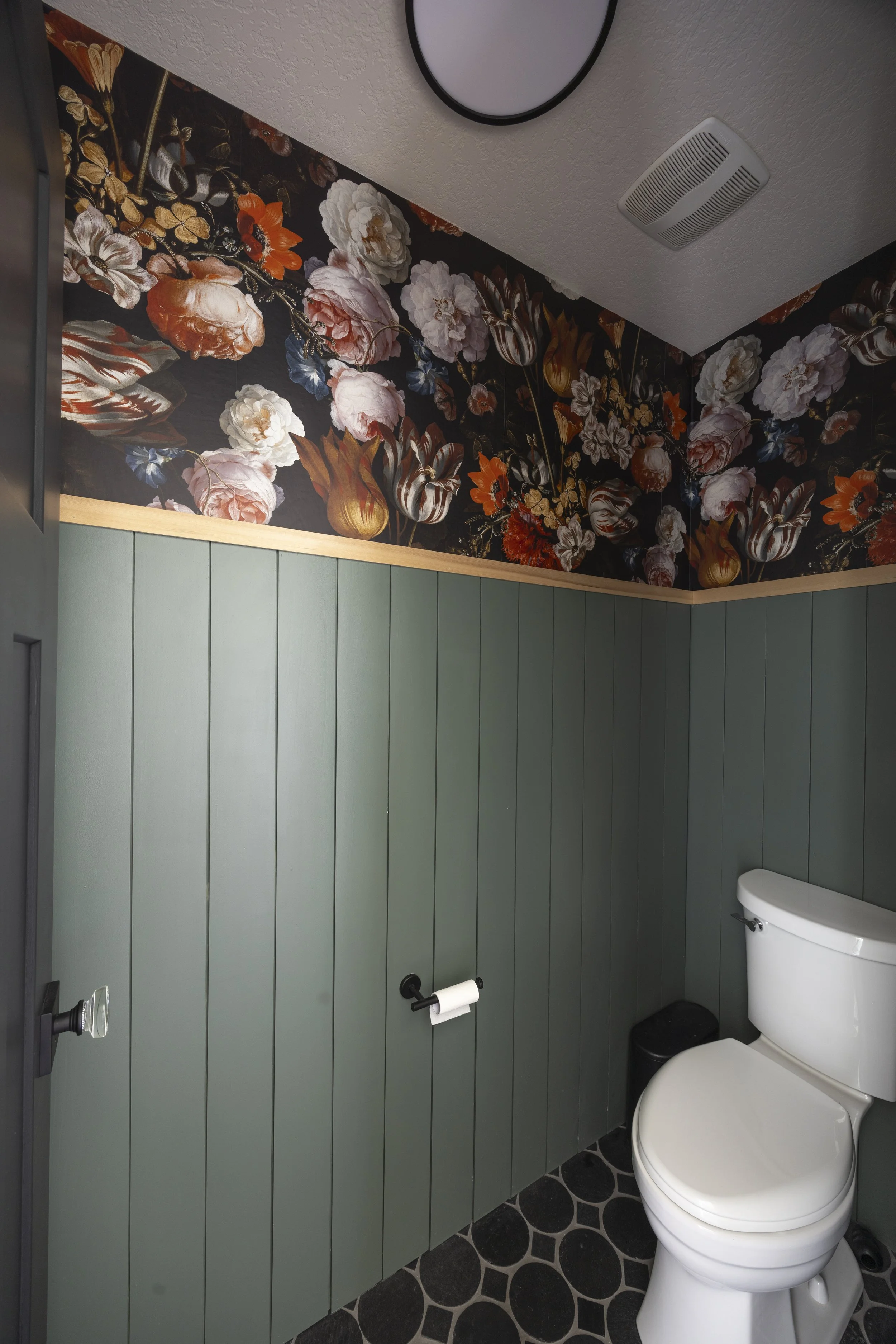 A bathroom with dark floral wallpaper above green vertical plank wainscoting and a wood ledge