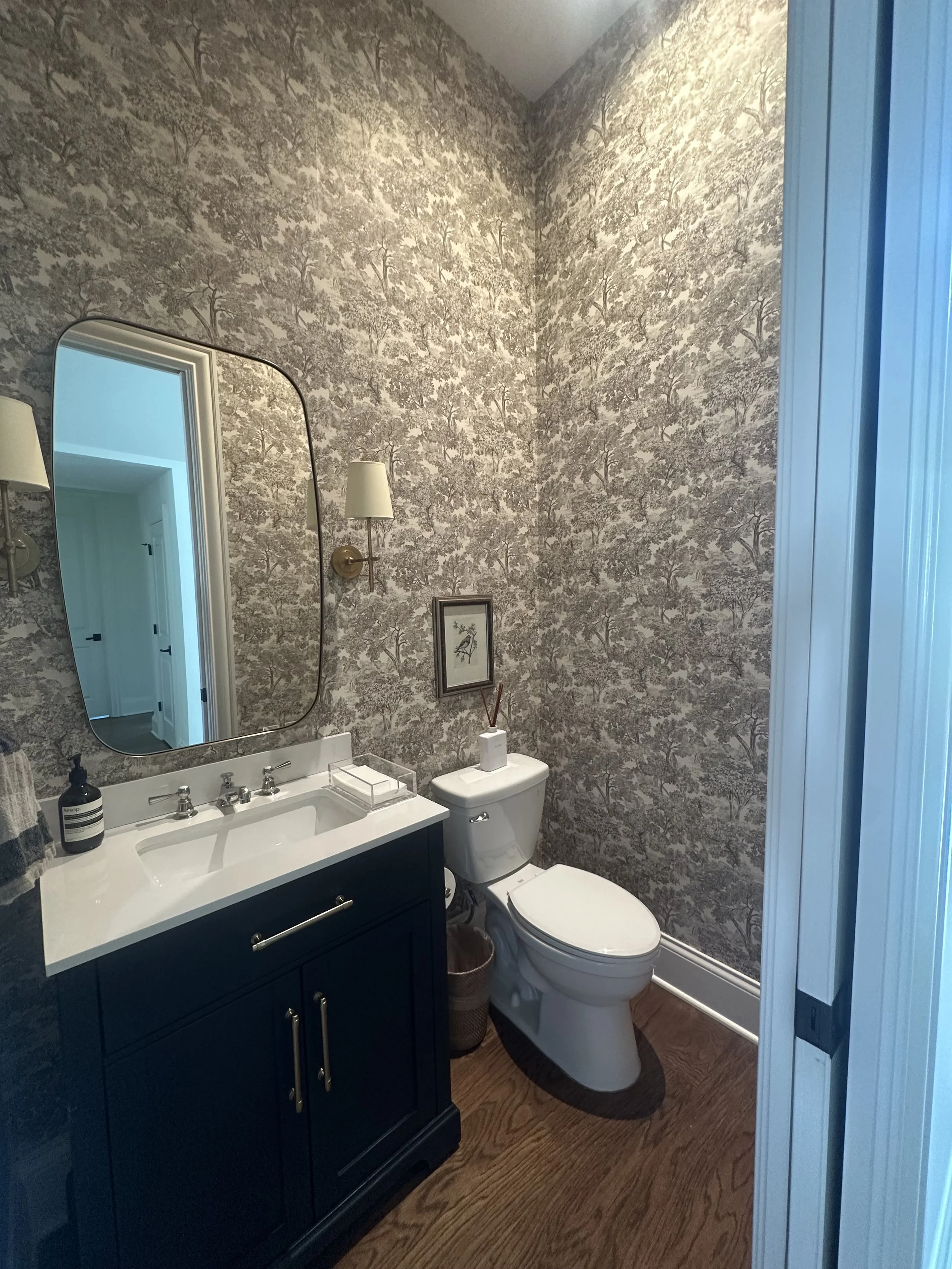 A powder room with grey botanical wallpaper, a navy vanity, a white counter, a brass-framed mirror, and a wall sconce.