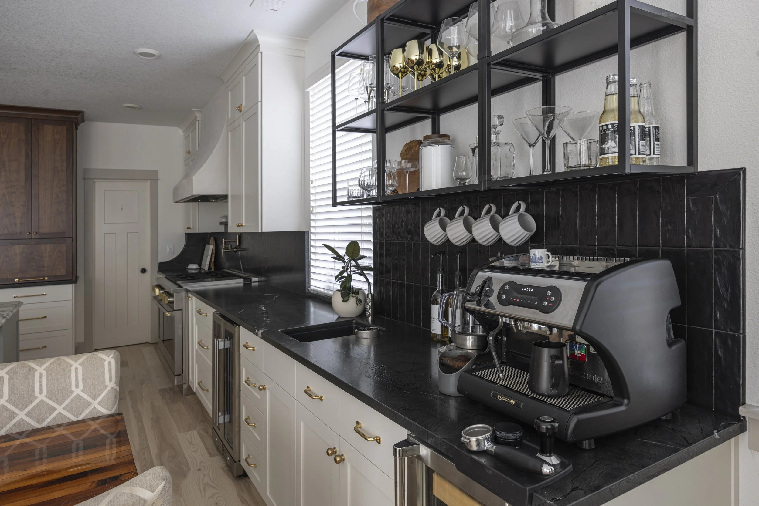 A kitchen coffee bar with white cabinets, a dark tile backsplash, open black shelving, and an espresso machine.