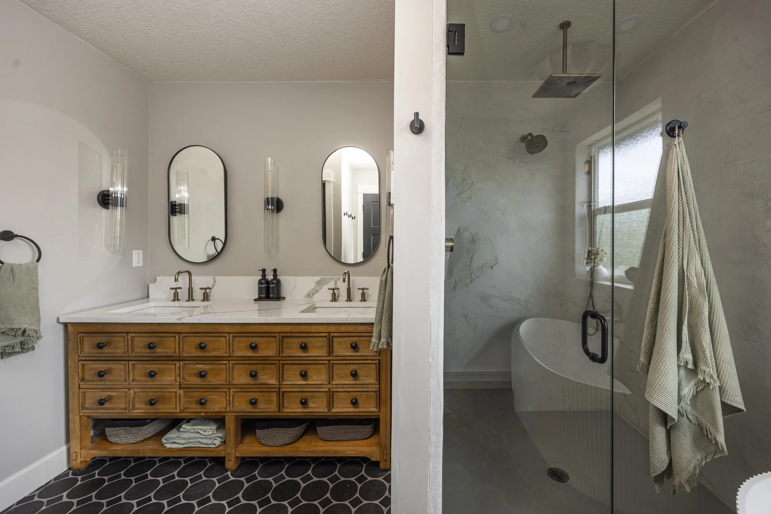 A master bathroom with a wood double vanity, white stone counters, black oval mirrors, a glass shower, a freestanding tub, and a black stone-tile floor.