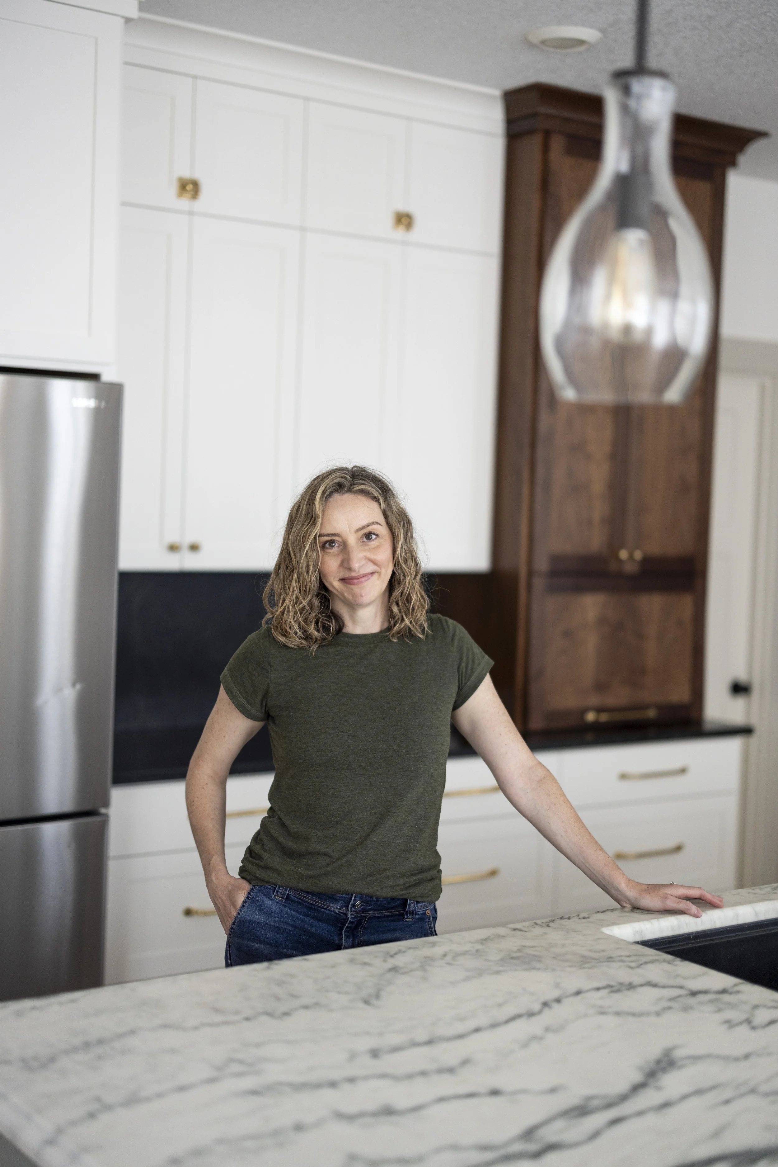 Tricia Caldwell, owner of Maven Made Home, standing in a renovated kitchen with white shaker cabinets and a tall walnut cabinet.