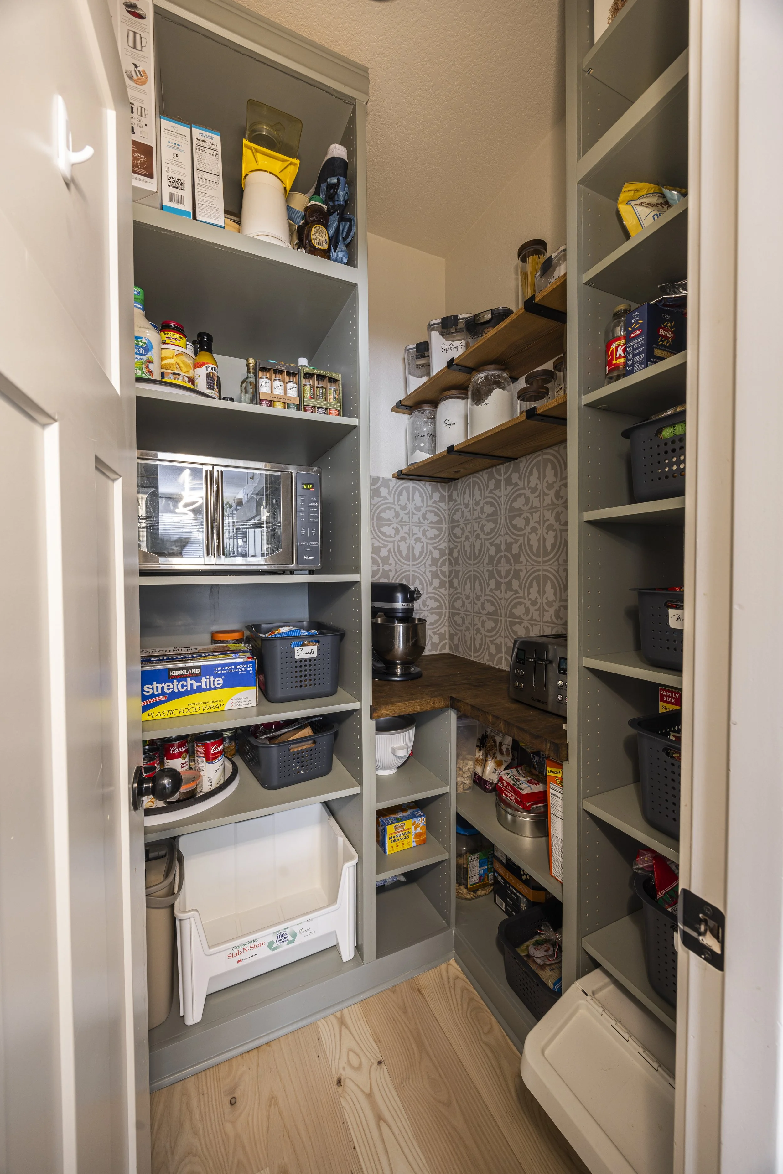 A walk-in pantry with grey-green built-in shelving, wood accent shelves, and stocked food storage