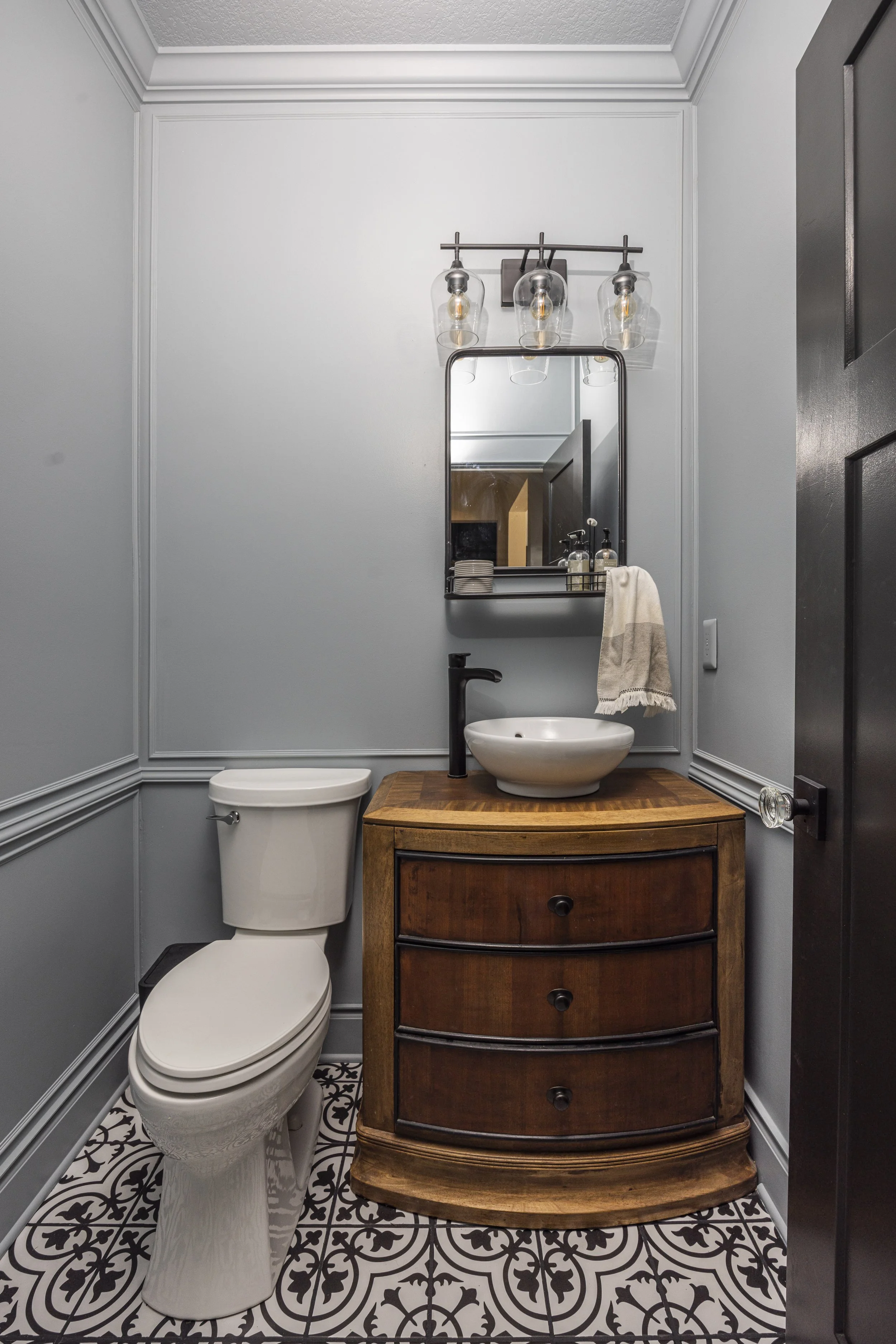 After renovation: A powder room with light gray walls and wainscoting, a wood vanity with a white vessel sink, a black faucet, and a black-framed mirror.